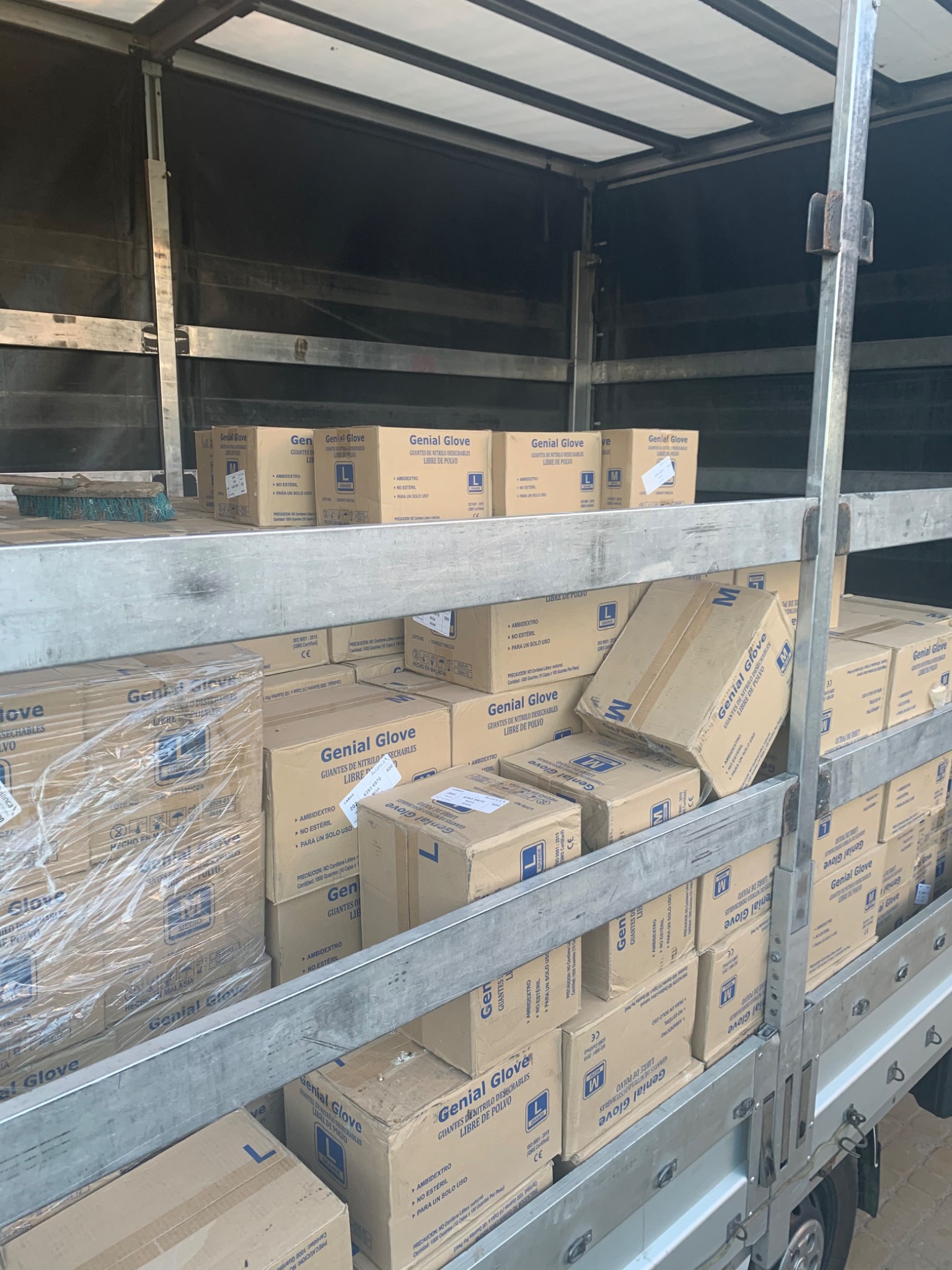 Wnętrze pojazdu dostawczego wypełnione kartonami rękawic nitrylowych marki Genial Glove, ułożonymi na metalowych półkach. Na górnej półce widoczna szczotka.