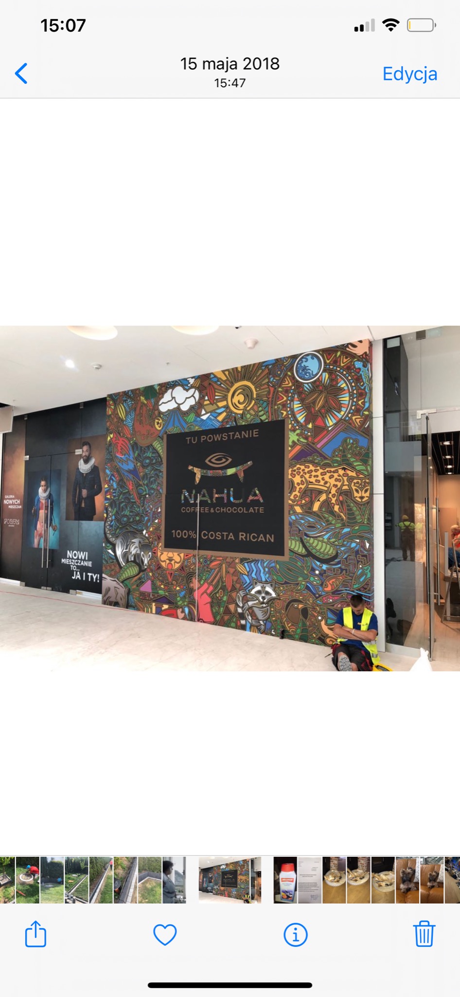 Kolorowa grafika ścienna z logo 'NAHLA COFFEE & CHOCOLATE 100% COSTA RICAN' w przestrzeni handlowej, obok witryny sklepowej i siedzącego pracownika w kamizelce odblaskowej.