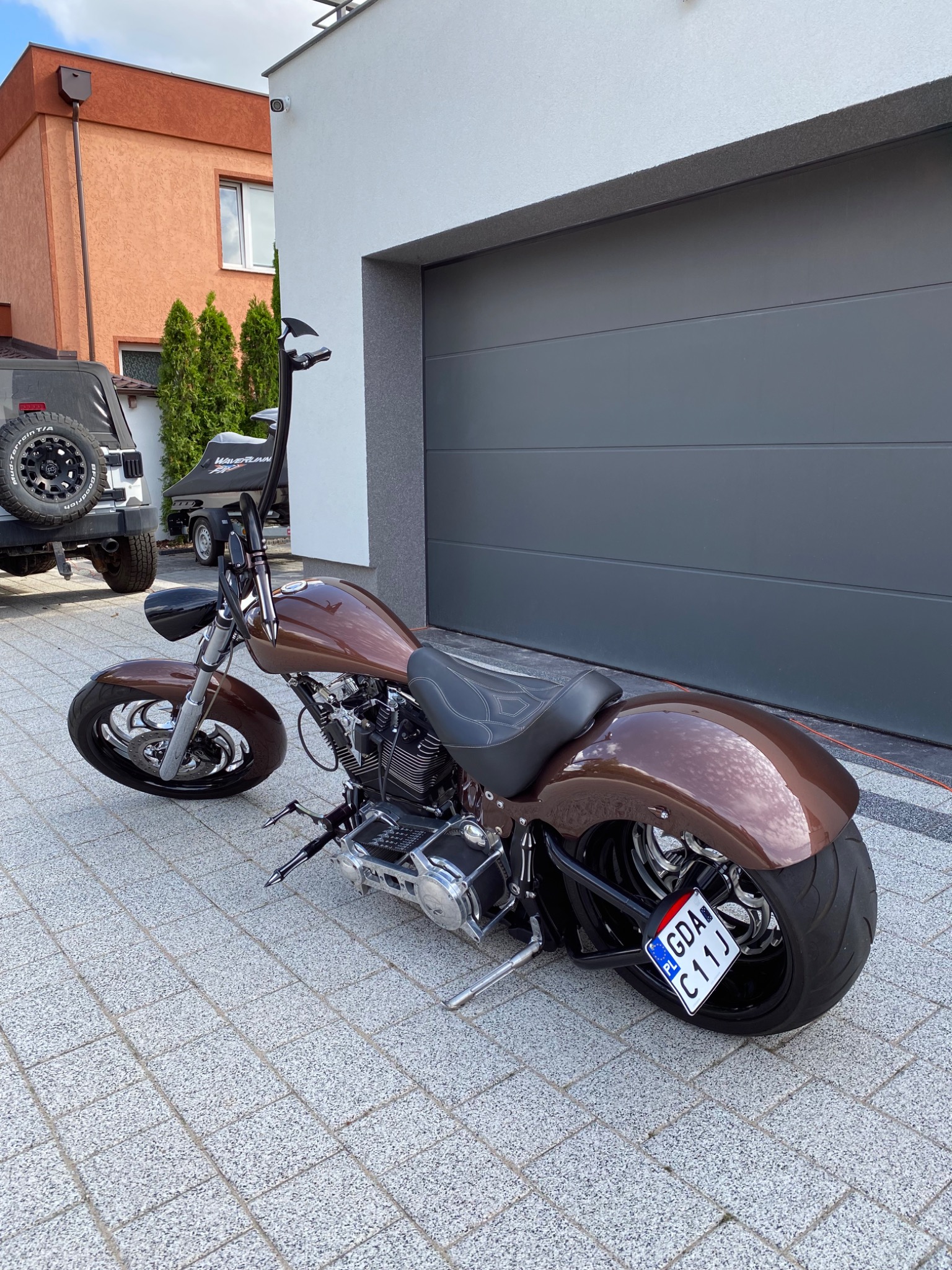 Brązowy motocykl typu chopper zaparkowany na kostce brukowej przed garażem, z widoczną tablicą rejestracyjną GDA C11J.