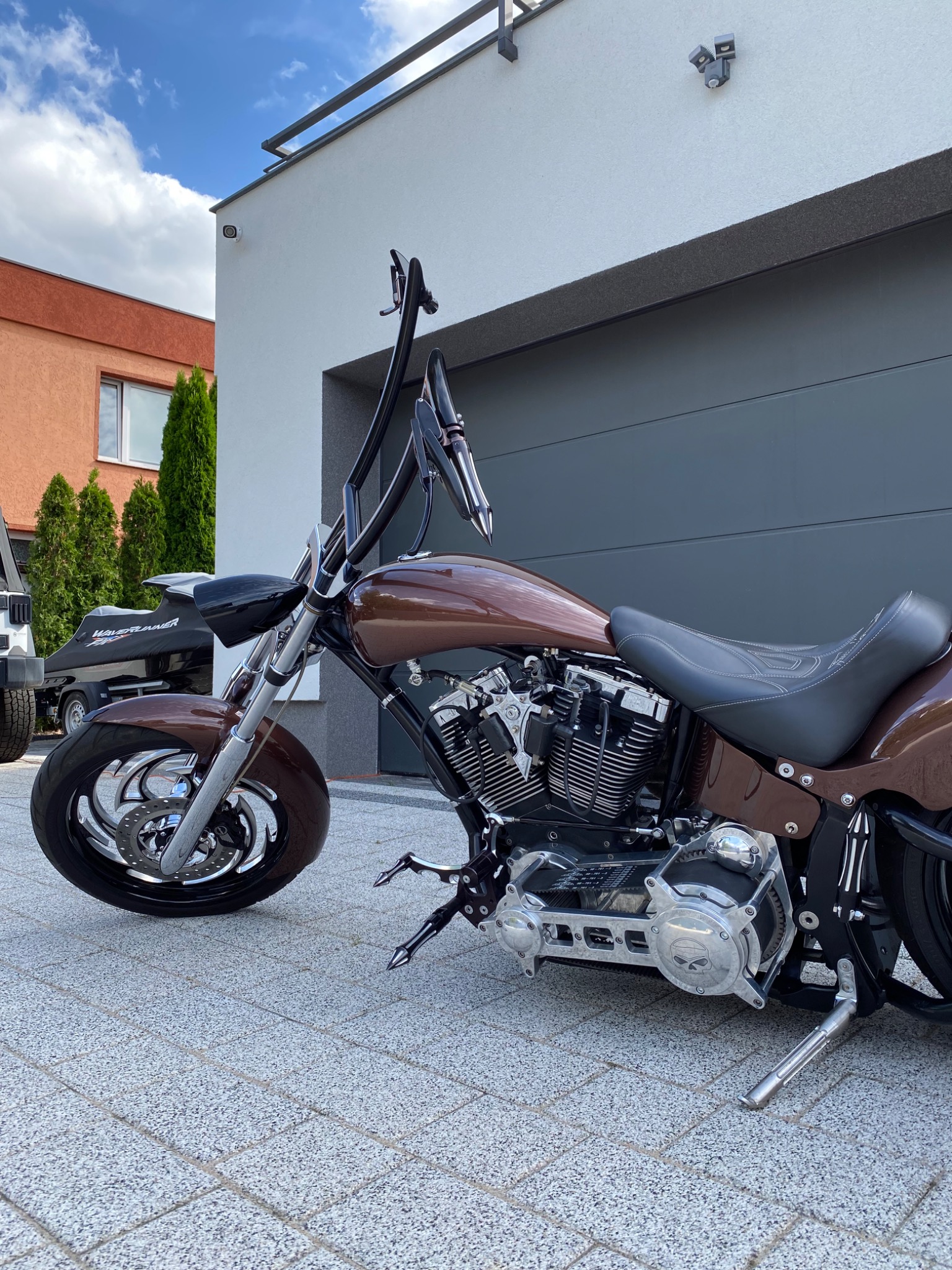 Brązowy motocykl typu chopper z chromowanymi elementami, zaparkowany na tle nowoczesnego domu z szarą bramą garażową i zielonymi tujami, w tle widać przyczepę do przewozu skutera wodnego.