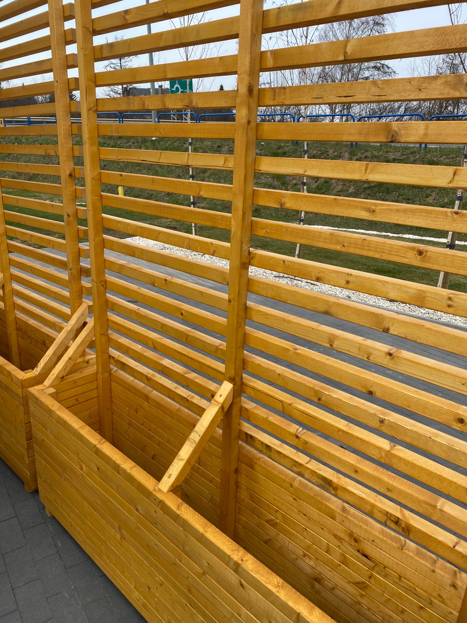 Drewniane, lakierowane donice z ażurową ścianką, ustawione wzdłuż drogi, z widocznymi znakami drogowymi w tle.