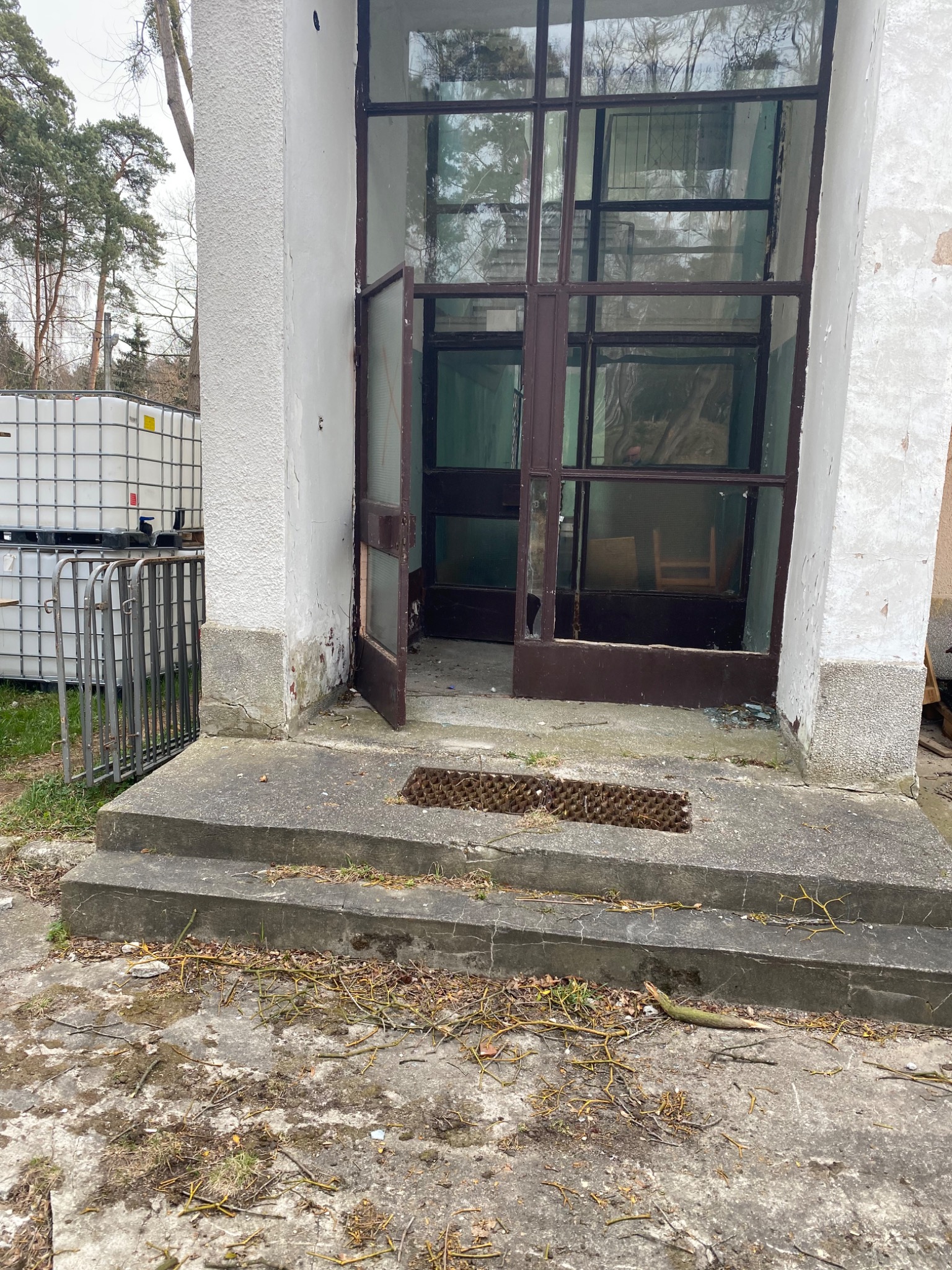 Zniszczone wejście do budynku z otwartymi, przeszklonymi drzwiami i trzema betonowymi stopniami, z widocznymi śladami zaniedbania i odpadającym tynkiem, na zewnątrz widoczne białe zbiorniki.