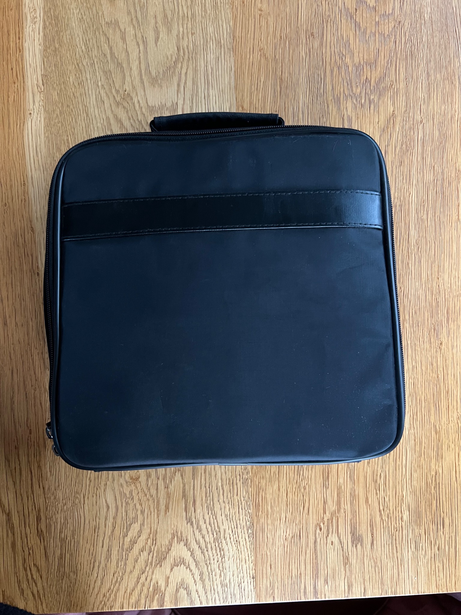 Zamknięta, czarna torba transportowa z uchwytem i zamkiem błyskawicznym, leżąca na drewnianym blacie.