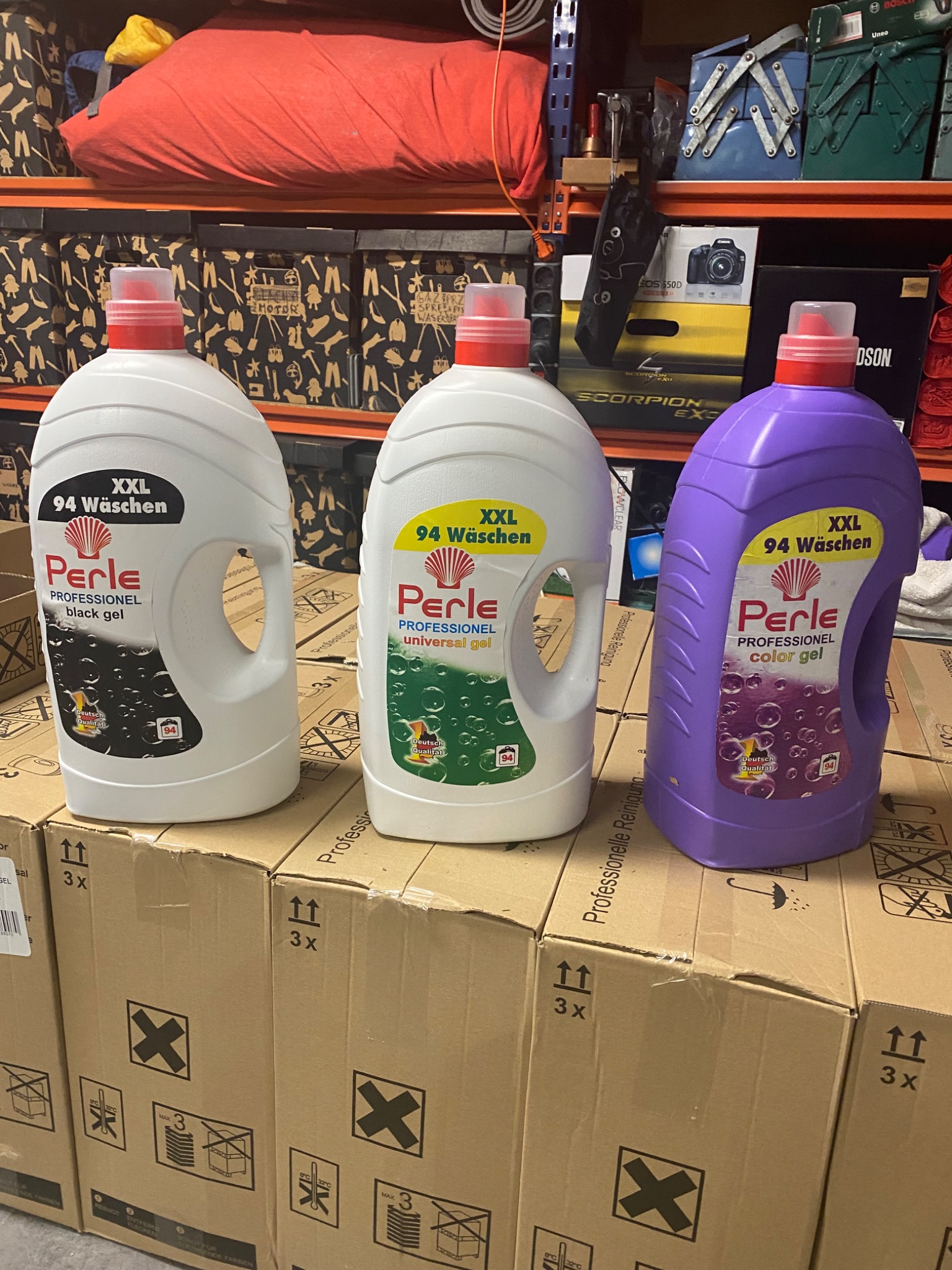 Trzy butelki profesjonalnego żelu do prania Perle (czarny, uniwersalny i kolorowy) stojące na kartonach w magazynie, widoczne symbole transportowe na kartonach.