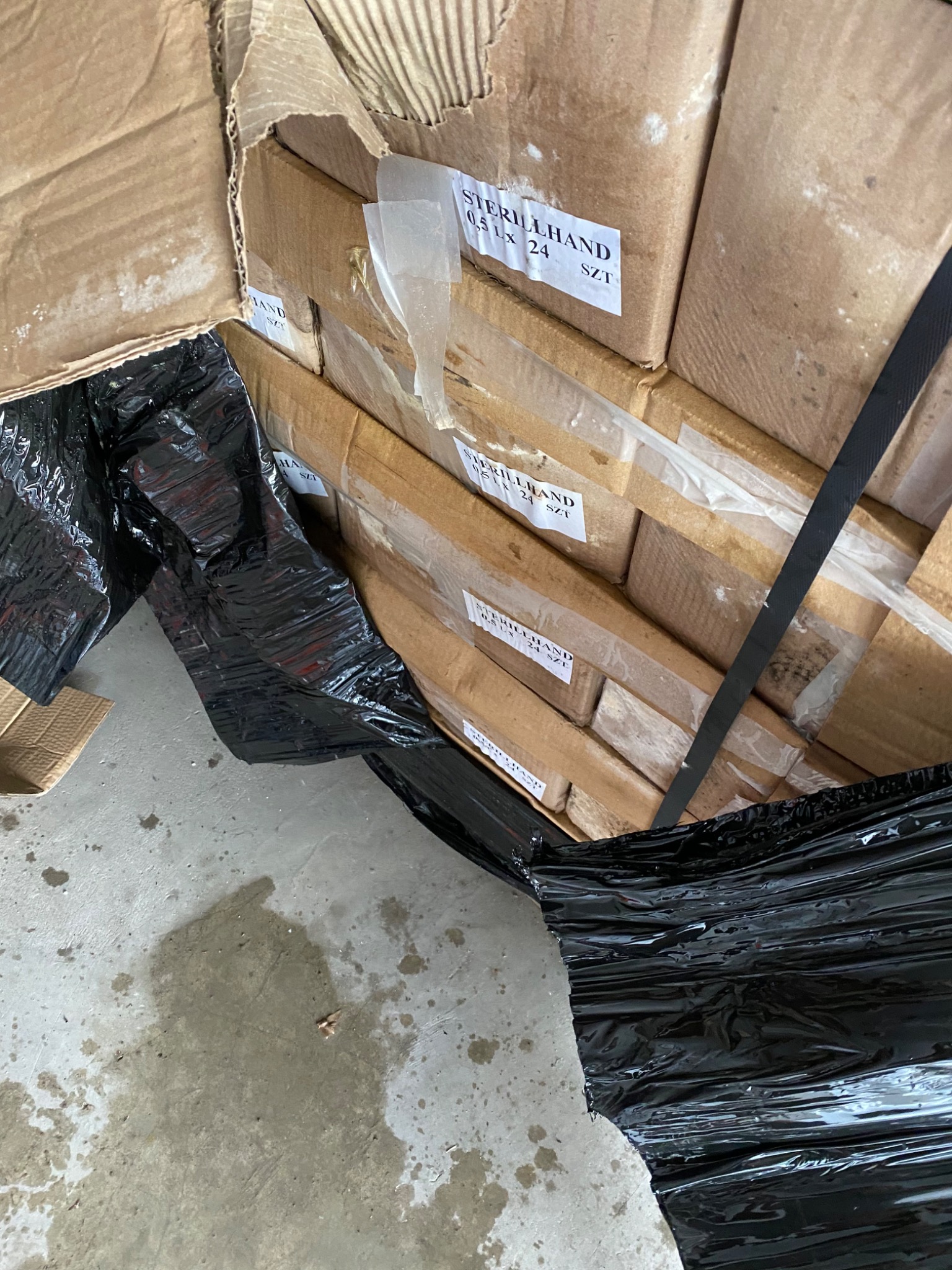 Kartonowe pudła z produktami Sterillhand 0,5 Lx 24 SZT, zabezpieczone czarną folią i pasami transportowymi, w otwartym kartonie zbiorczym.