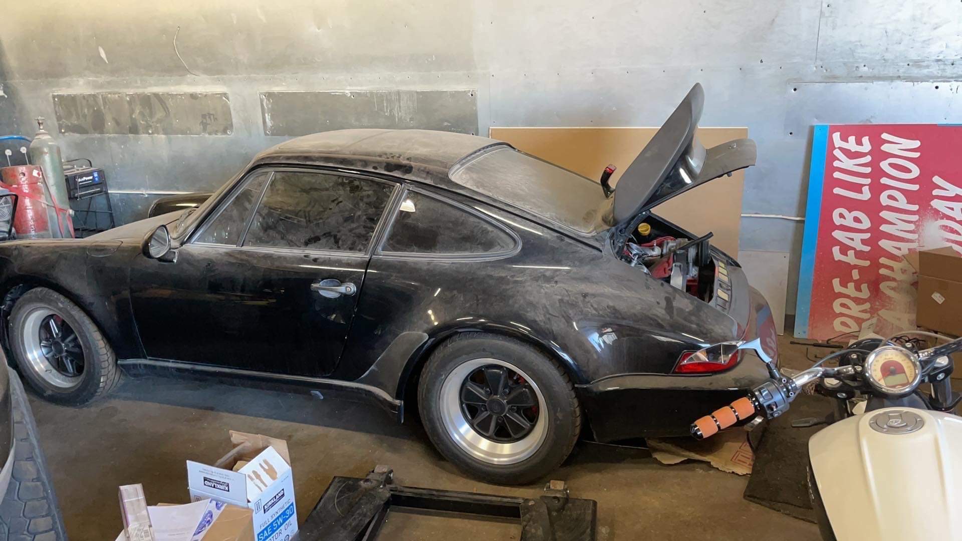 Zakurzony czarny samochód sportowy marki Porsche z otwartym bagażnikiem, stojący w garażu w Gdańsku, obok motocykla i kartonów z olejem silnikowym.