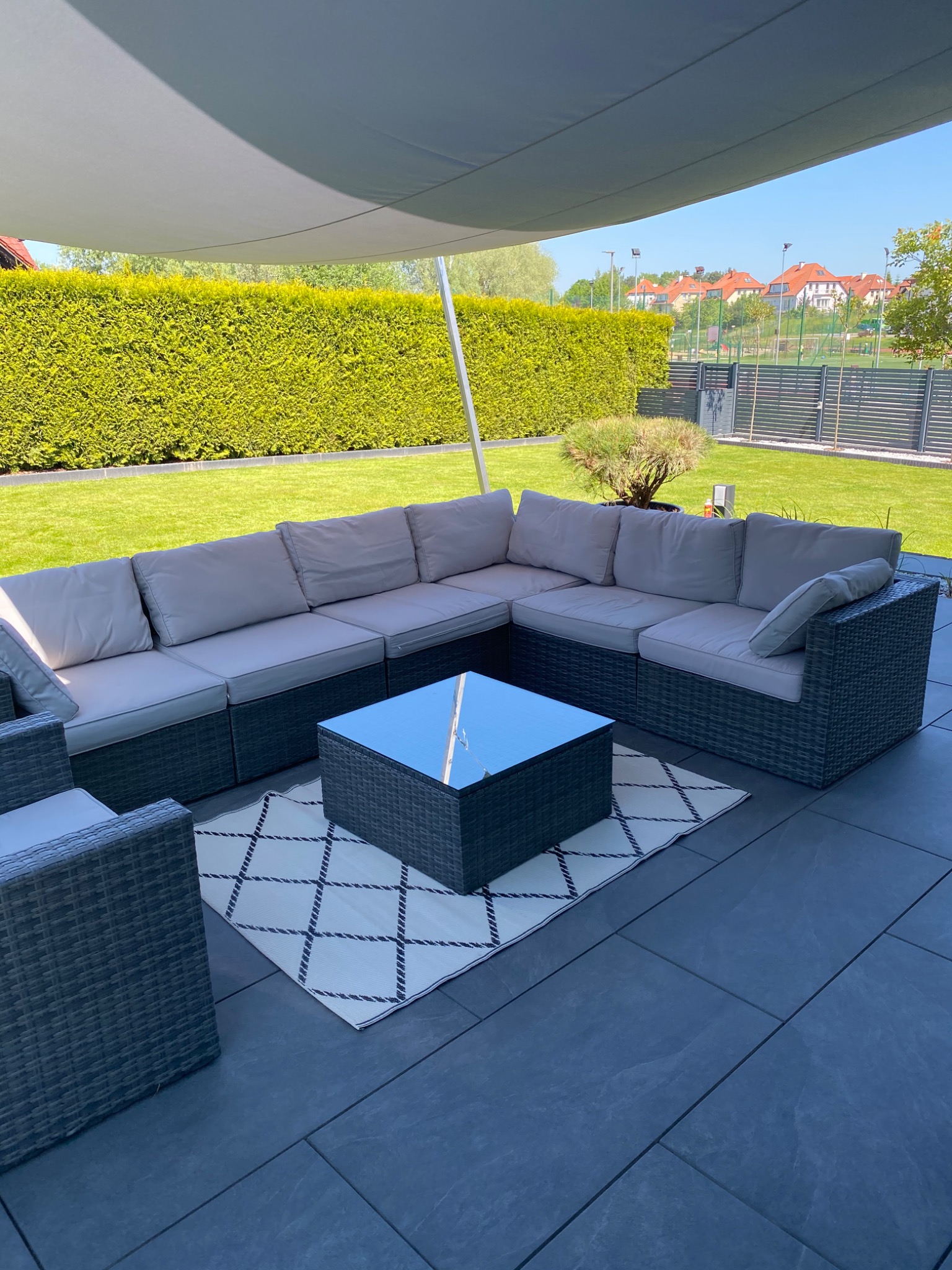 Zadaszony taras z szarą, modułową sofą rattanową z jasnymi poduszkami, szklanym stolikiem kawowym i dywanem w geometryczne wzory, na tle zadbanego trawnika i żywopłotu.