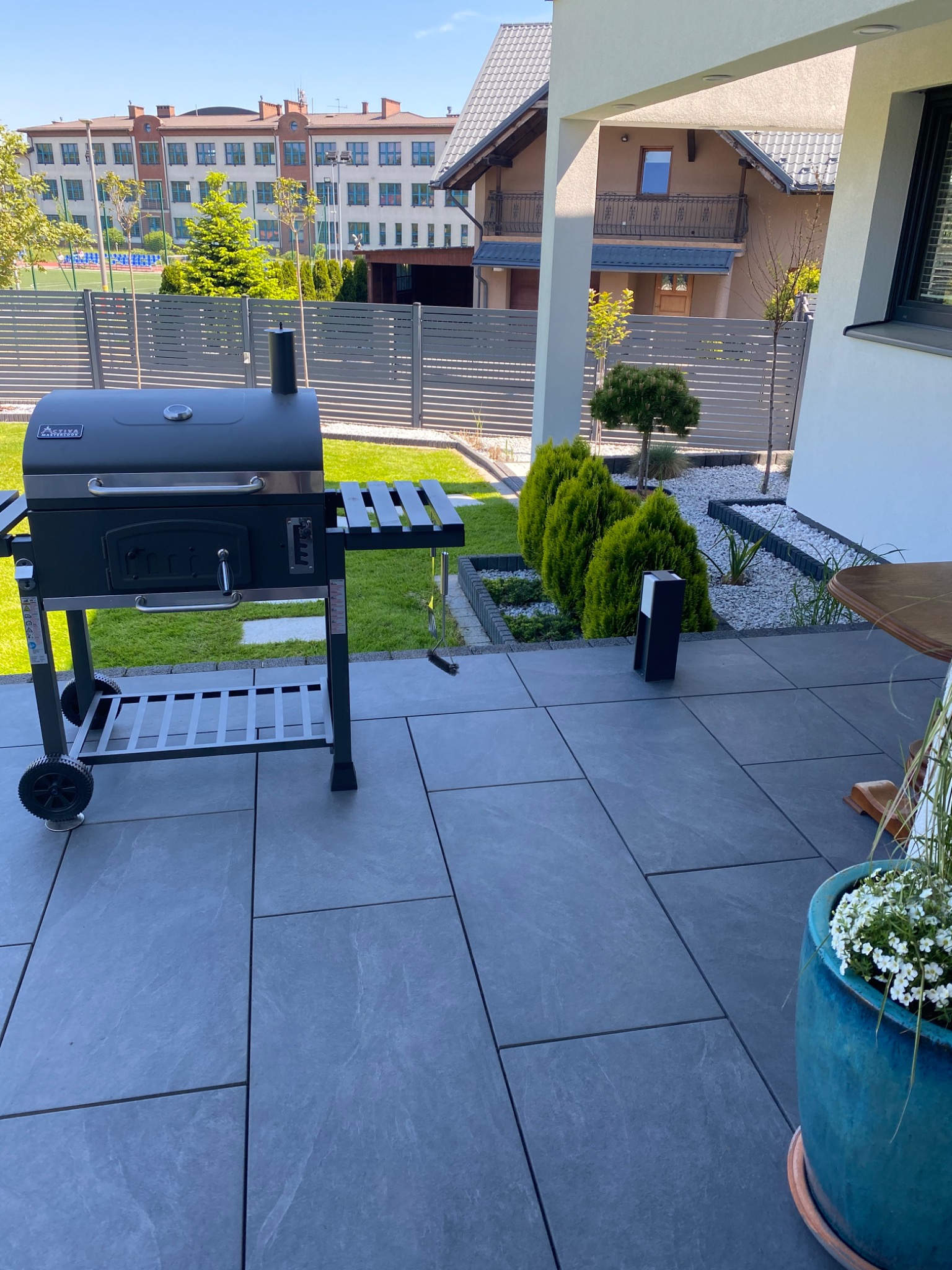 Nowoczesny grill węglowy z wędzarnią marki Activa na tarasie z szarych płyt, w tle zadbany trawnik i budynki mieszkalne.