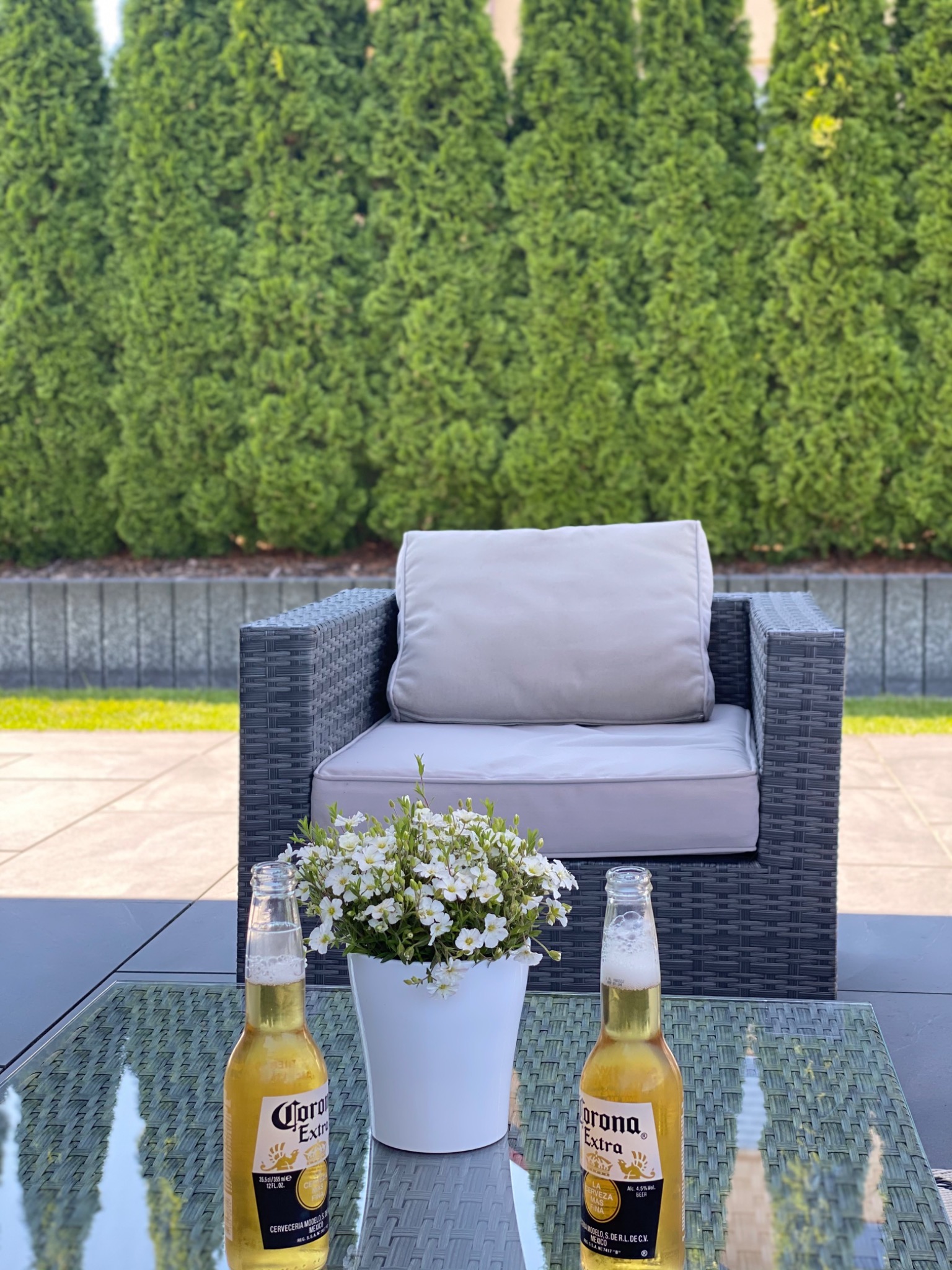 Relaksujący kącik na tarasie: rattanowy fotel z poduszkami, stolik kawowy z dwoma butelkami piwa Corona i doniczką z białymi kwiatami na tle ściany z tuj.