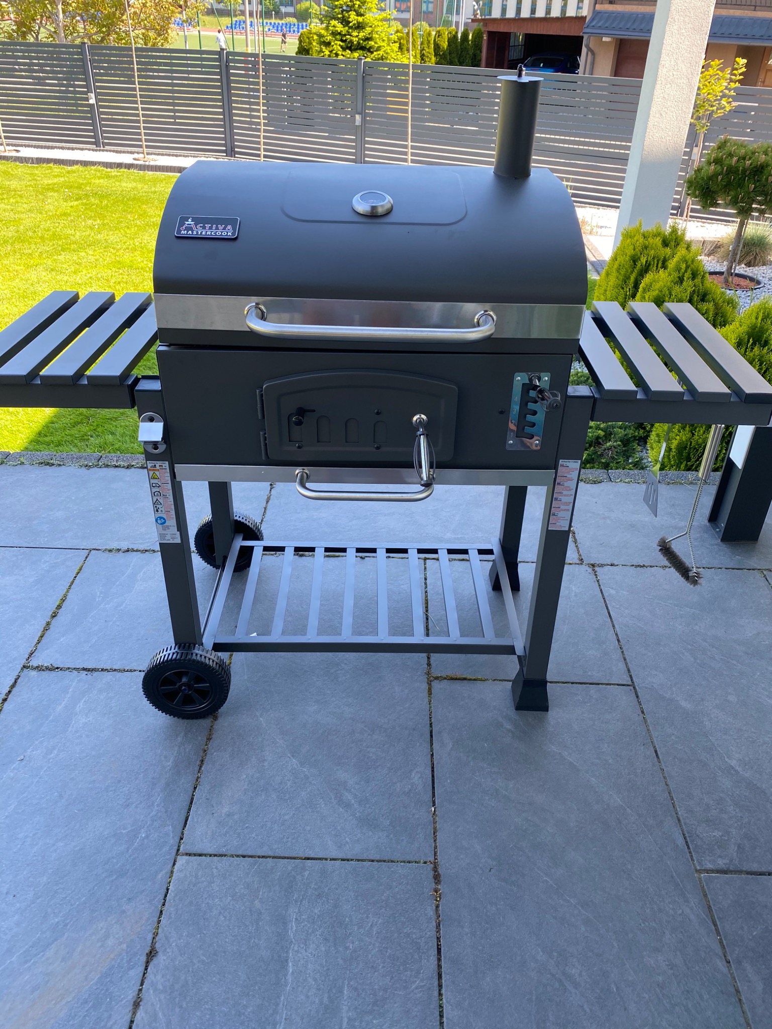 Nowoczesny, czarny grill węglowy z bocznymi półkami na kółkach, stojący na szarym tarasie z płyt kamiennych w ogrodzie.