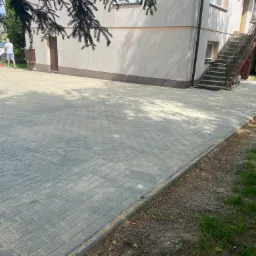 Szara kostka brukowa ułożona przed budynkiem z zewnętrznymi schodami; widoczny mężczyzna zamiatający powierzchnię.