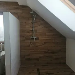 Ukośna łazienka na poddaszu z drewnopodobnymi płytkami na ścianach i podłodze, kabina prysznicowa bez brodzika z odpływem liniowym i oknem dachowym.