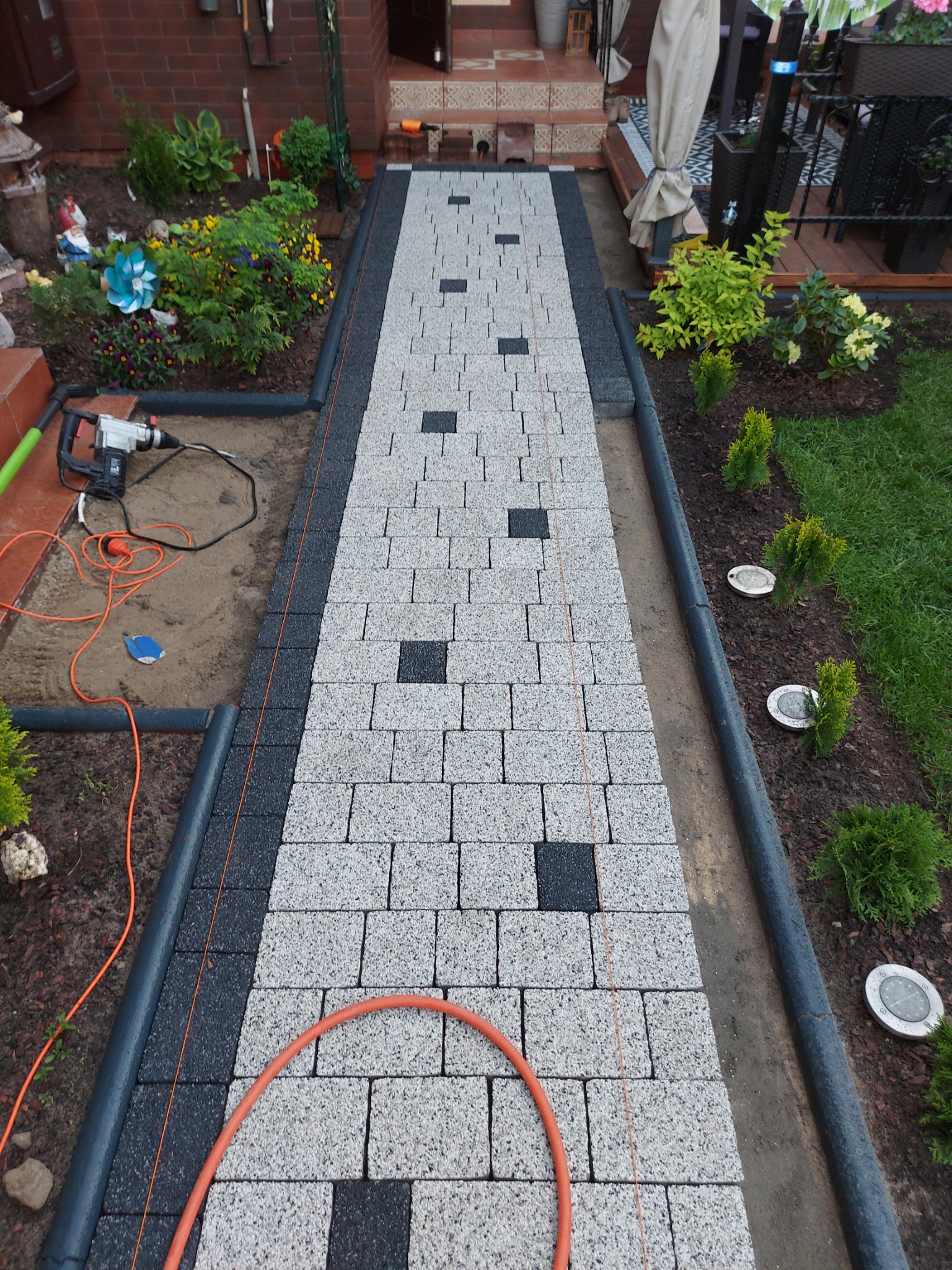 Ścieżka z jasnoszarych kostek brukowych z czarnymi akcentami, obrzeża z ciemnej kostki, widoczne narzędzia i pomarańczowy wąż, ogród z roślinami i lampami solarnymi po bokach.