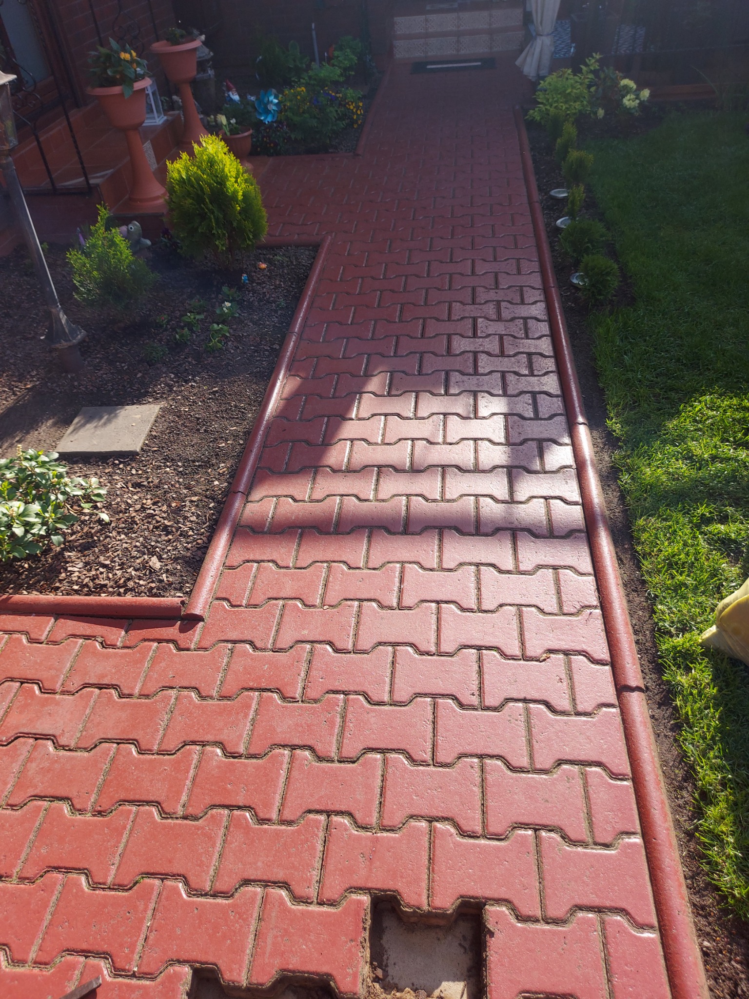 Czerwony chodnik z kostki brukowej w kształcie litery H, z widocznym brakiem jednej kostki i ziemią w dziurze, obrzeża chodnika w kolorze ceglastym, obok trawnik i rabaty z roślinami.