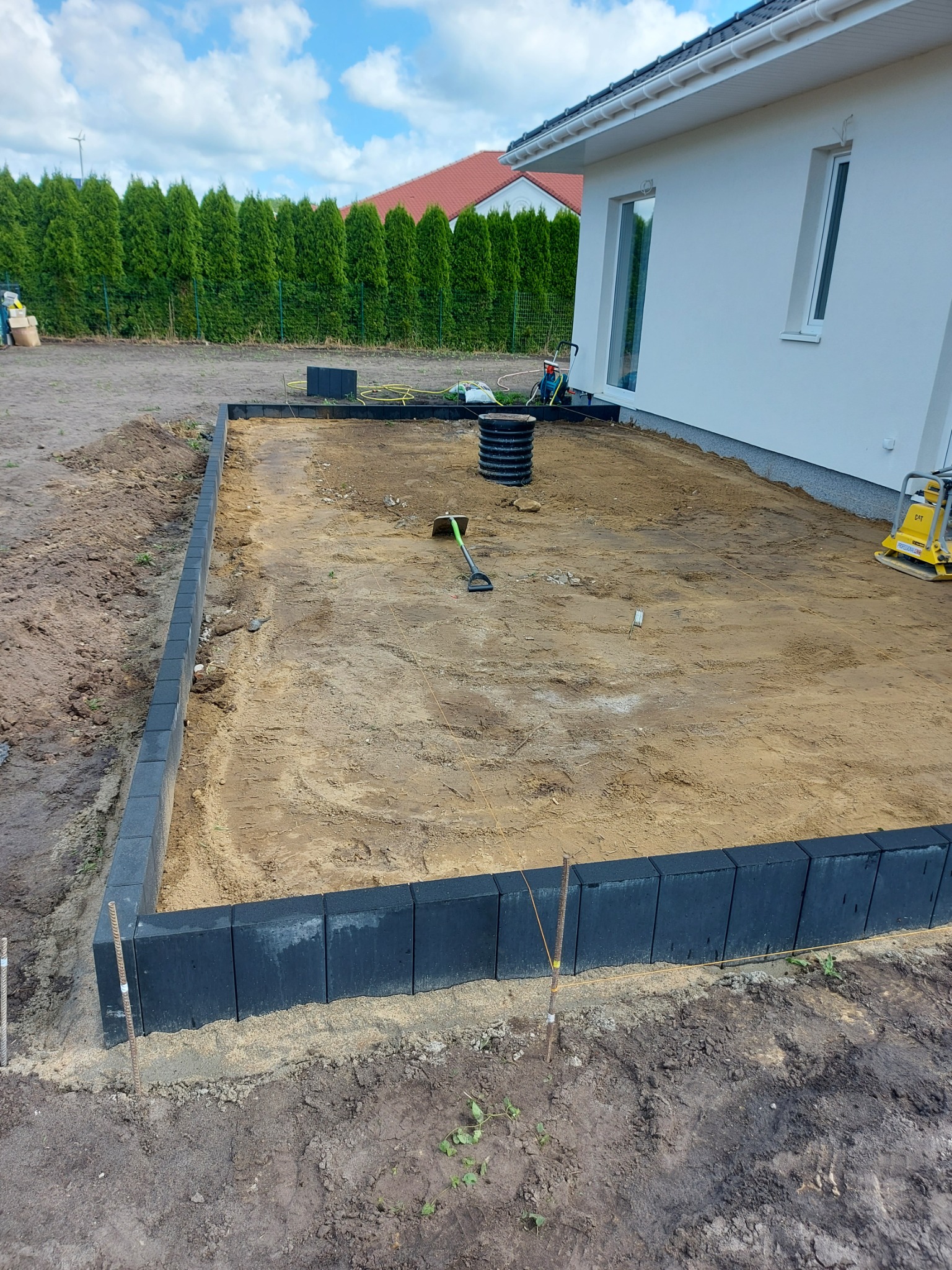 Przygotowanie terenu pod taras: wyrównana piaskiem powierzchnia ograniczona czarnymi obrzeżami z kostki, widoczna ubijarka w tle, obok biały budynek z oknami.