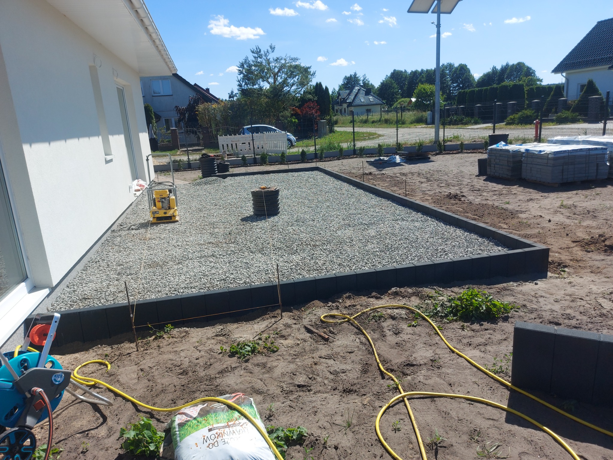 Przygotowanie podłoża pod taras: wysypany żwir, obrzeża z ciemnej kostki, w tle dom jednorodzinny i niebo z chmurami.