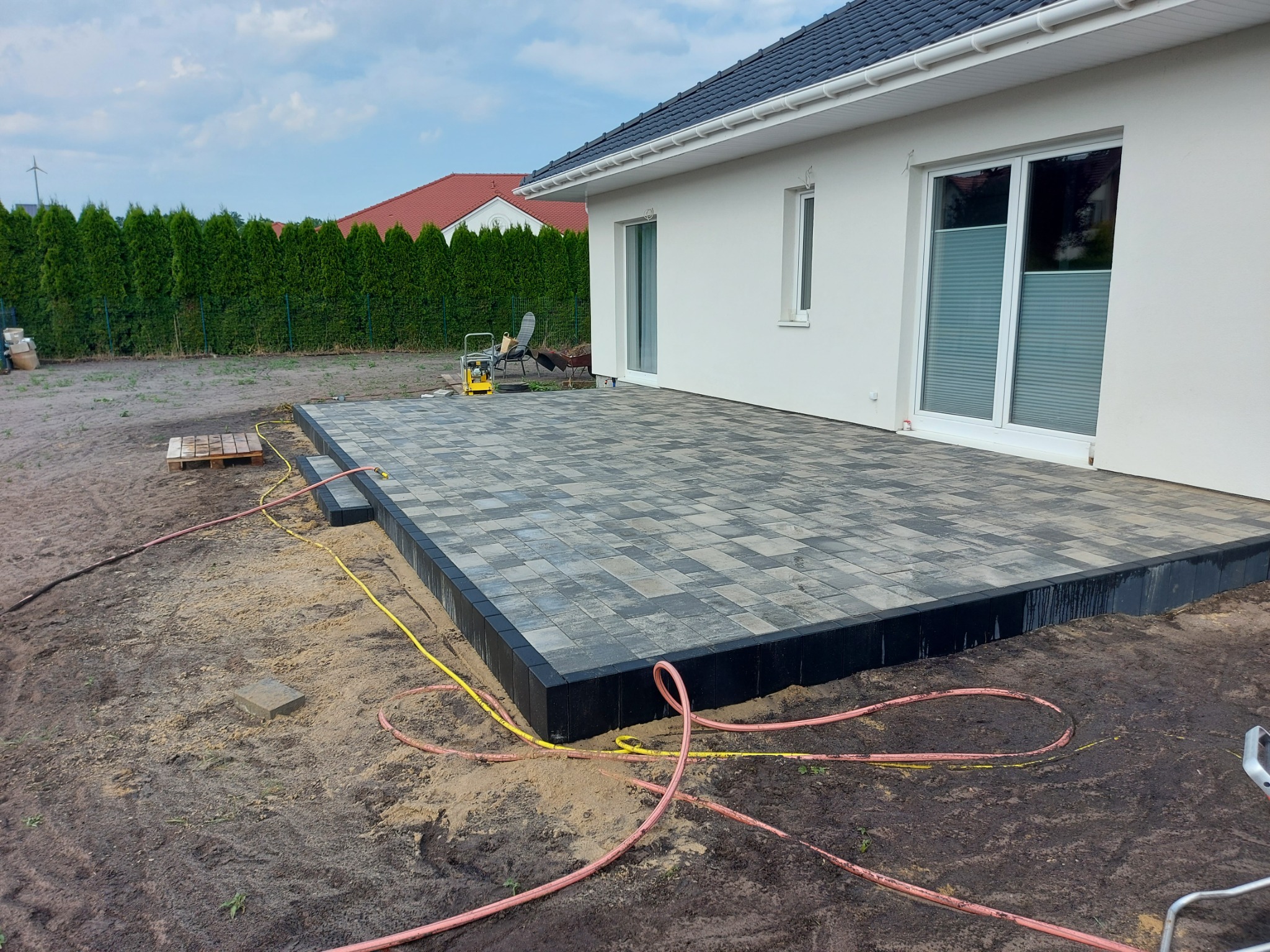 Nowo ułożony taras z kostki brukowej w szaro-czarnych odcieniach, z widocznymi śladami wilgoci na obrzeżach i ziemi, żółty wąż ogrodowy leżący na ziemi, w tle biały dom z ciemnym dachem i zielone...