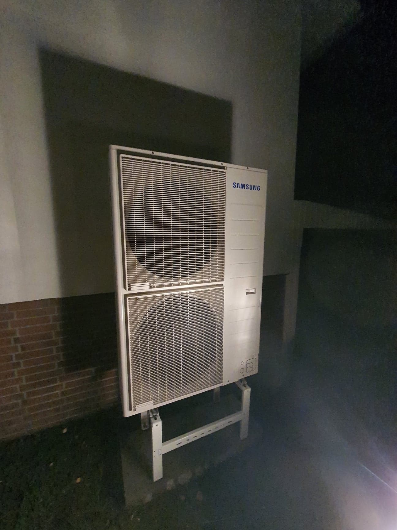 Zewnętrzna jednostka klimatyzacyjna Samsung, dwu-wentylatorowa, zamontowana na wsporniku, oświetlona z dołu, z widocznym cieniem na ścianie budynku.