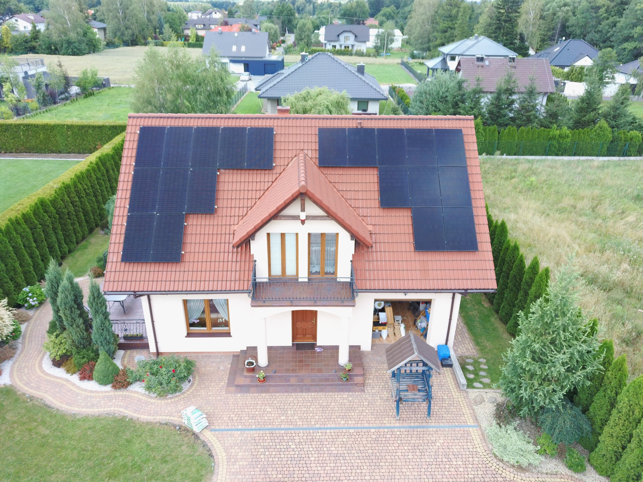 Widok z góry na dom jednorodzinny z panelami słonecznymi na dachu o ceglastym kolorze, otoczony zielonym ogrodem i innymi zabudowaniami.