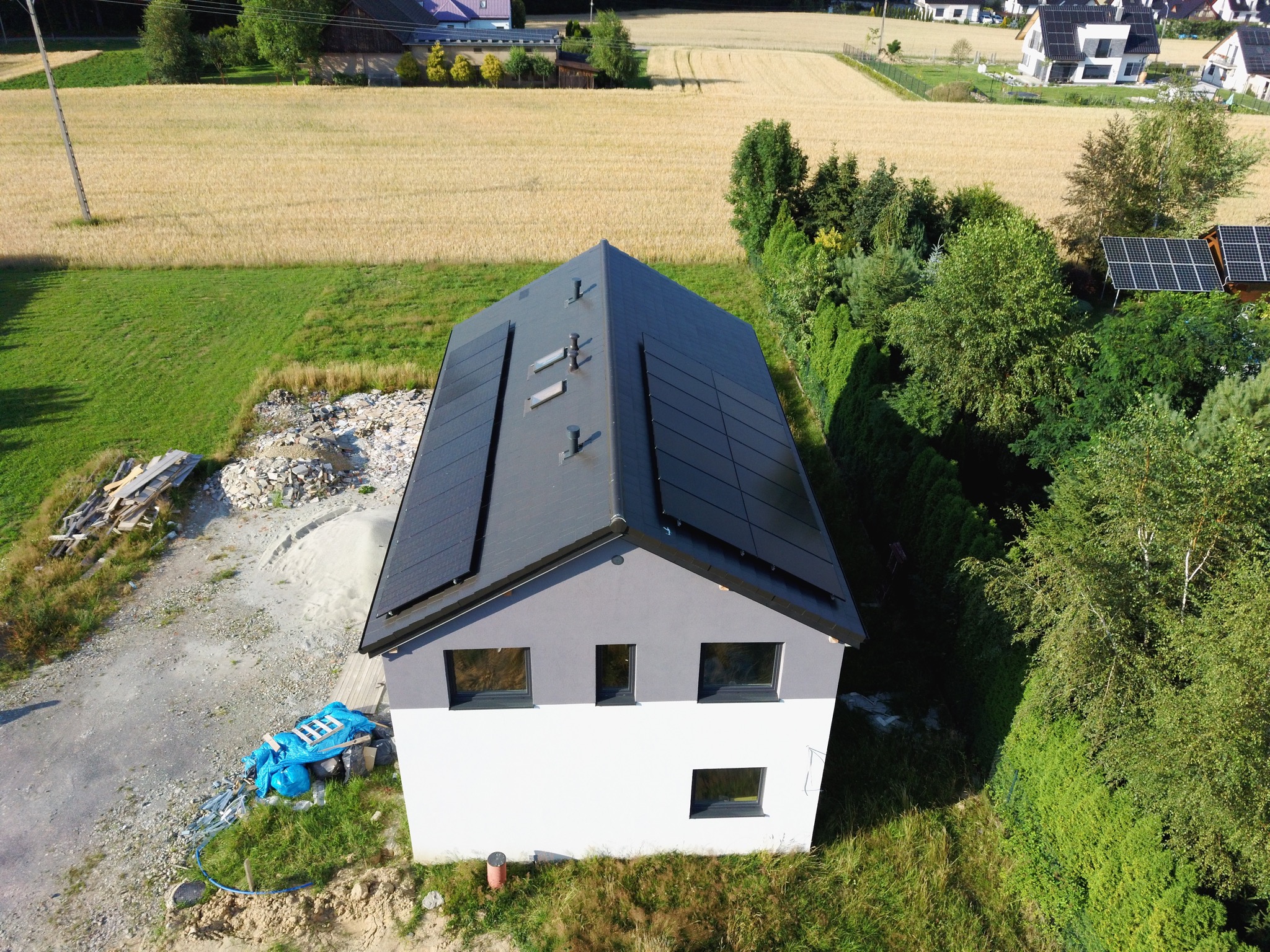 Widok z góry na nowoczesny dom z panelami słonecznymi na dachu, obok plac budowy z gruzem i materiałami budowlanymi, w tle pole zboża i inne domy.