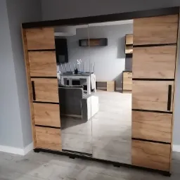 Nowoczesna szafa przesuwna z lustrami, wykończona okleiną drewnopodobną w odcieniach brązu, odbijająca fragment salonu z szarą sofą i stołem z krzesłami.