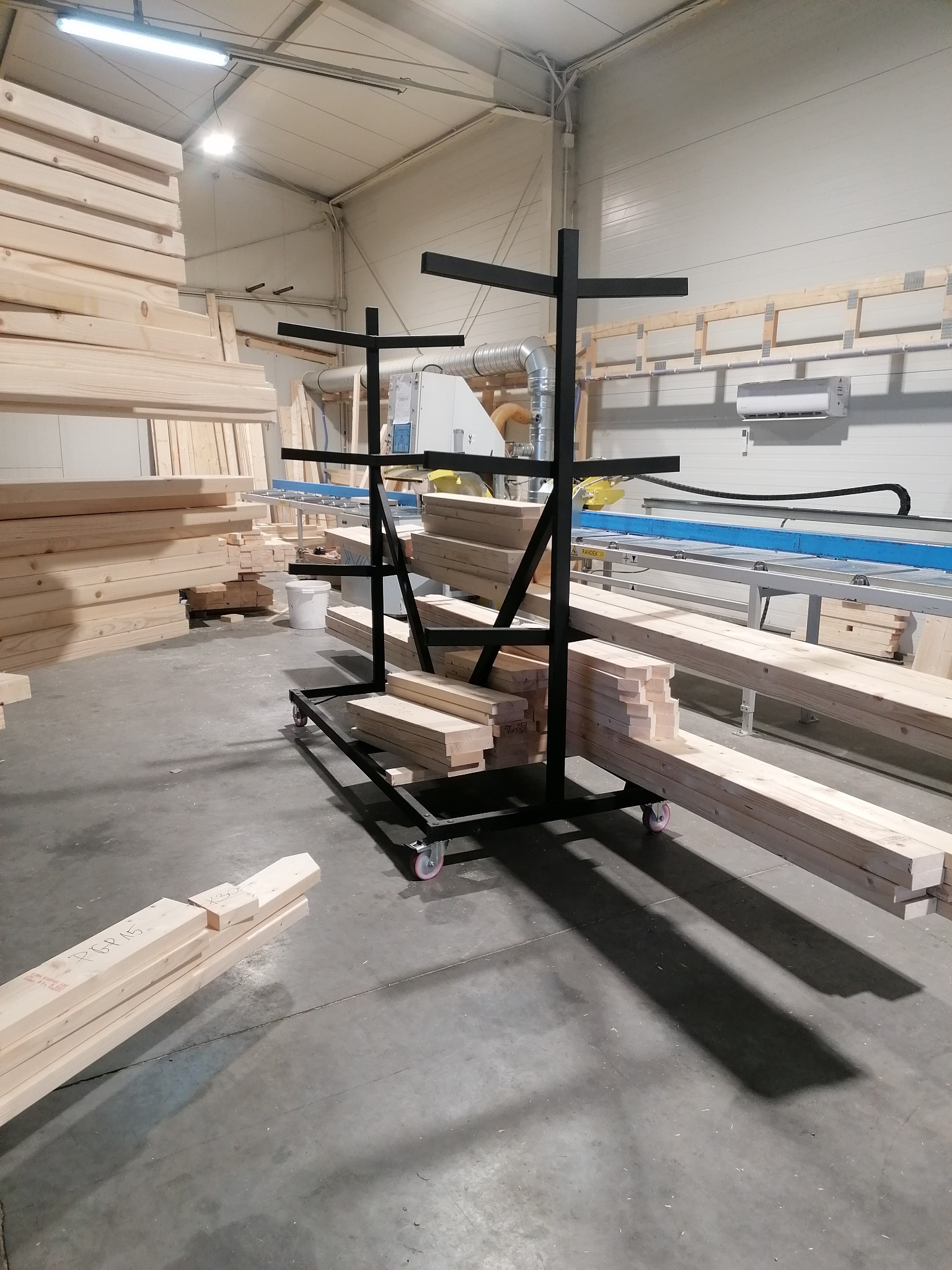 Stolarz w warsztacie: drewniane belki składowane na czarnym wózku transportowym z kołami, w tle maszyny stolarskie i stosy drewna.