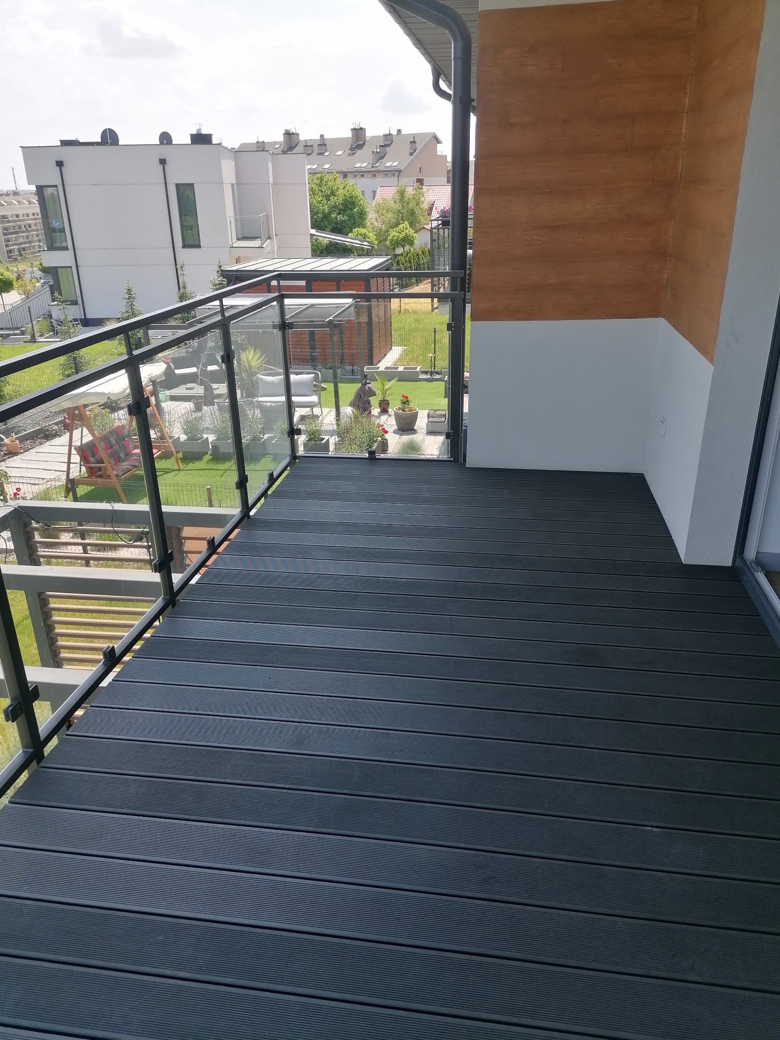 Nowoczesny balkon z czarną deską kompozytową, szklaną balustradą i widokiem na zadbany ogród z huśtawką i roślinami w donicach.