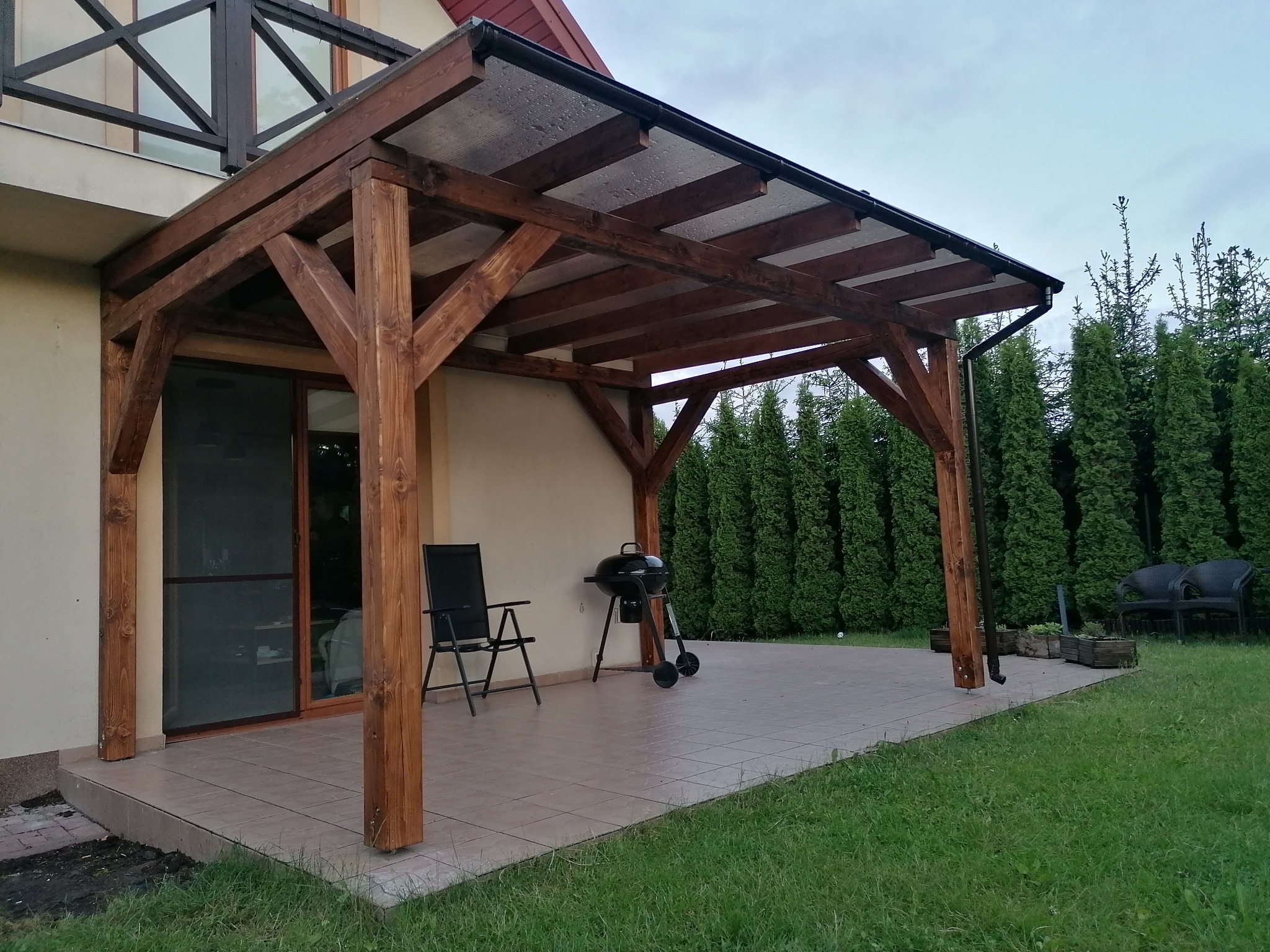 Drewniana pergola z przezroczystym zadaszeniem przylegająca do domu, z grillem i krzesłem na tarasie wyłożonym płytkami.