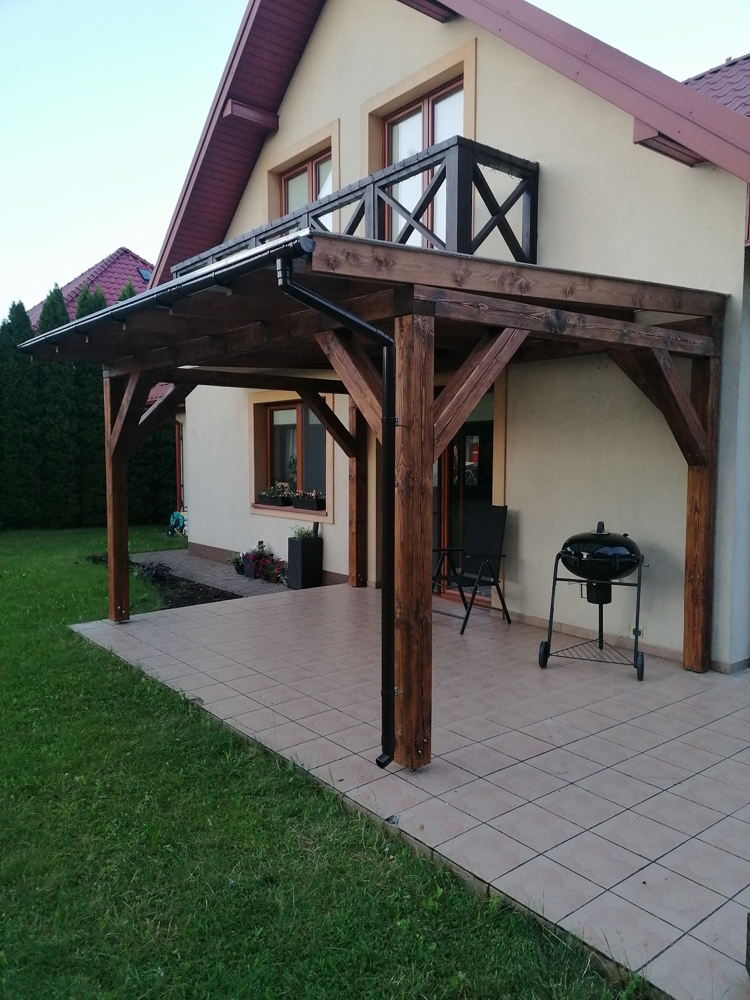 Drewniana wiata przydomowa z ciemnobrązowymi belkami, widok na elewację domu z balkonem, na tarasie grill.