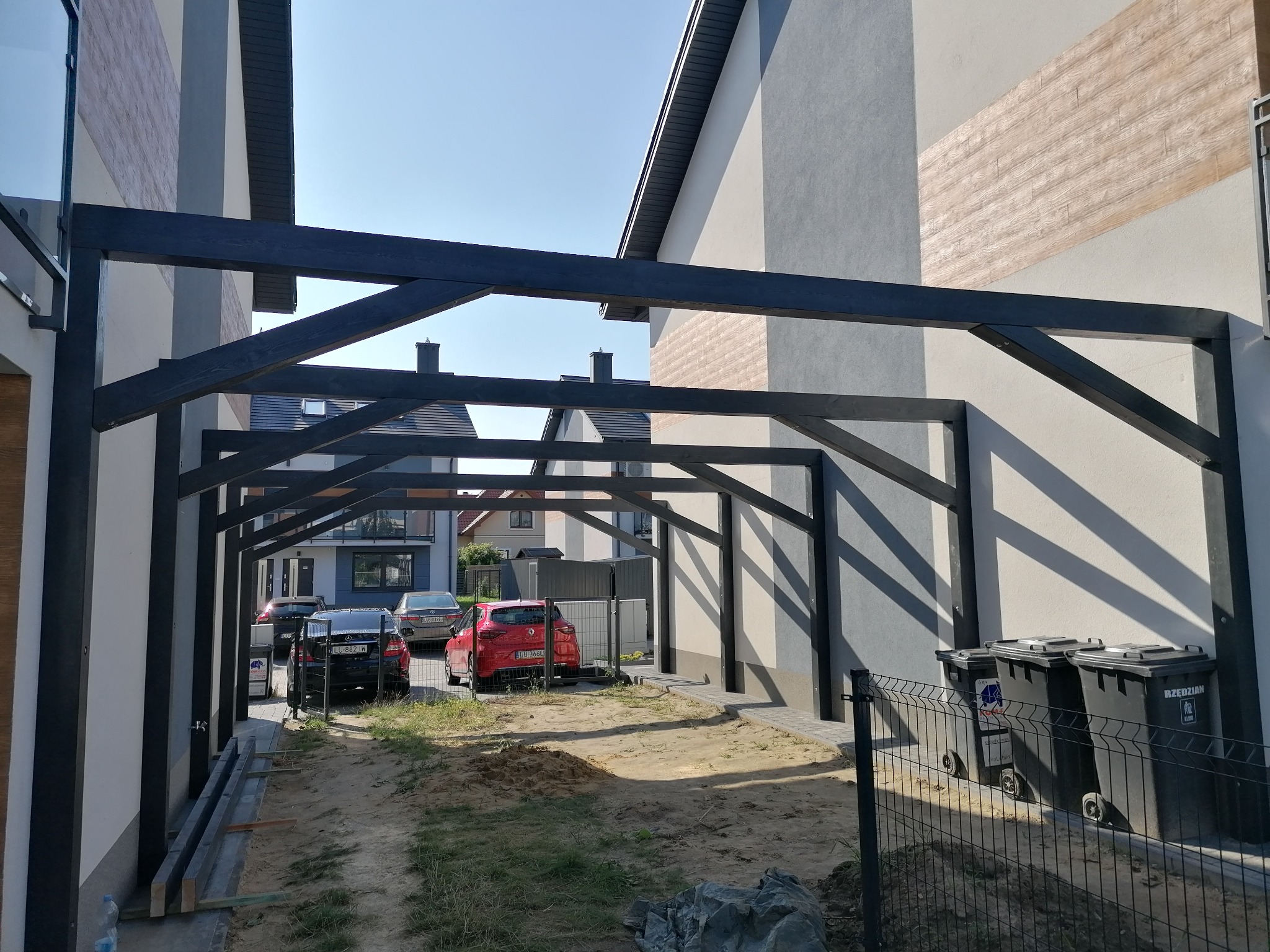 Konstrukcja z ciemnego drewna tworząca zadaszenie nad miejscem parkingowym, widoczne samochody i domy w tle, ziemia nieutwardzona.