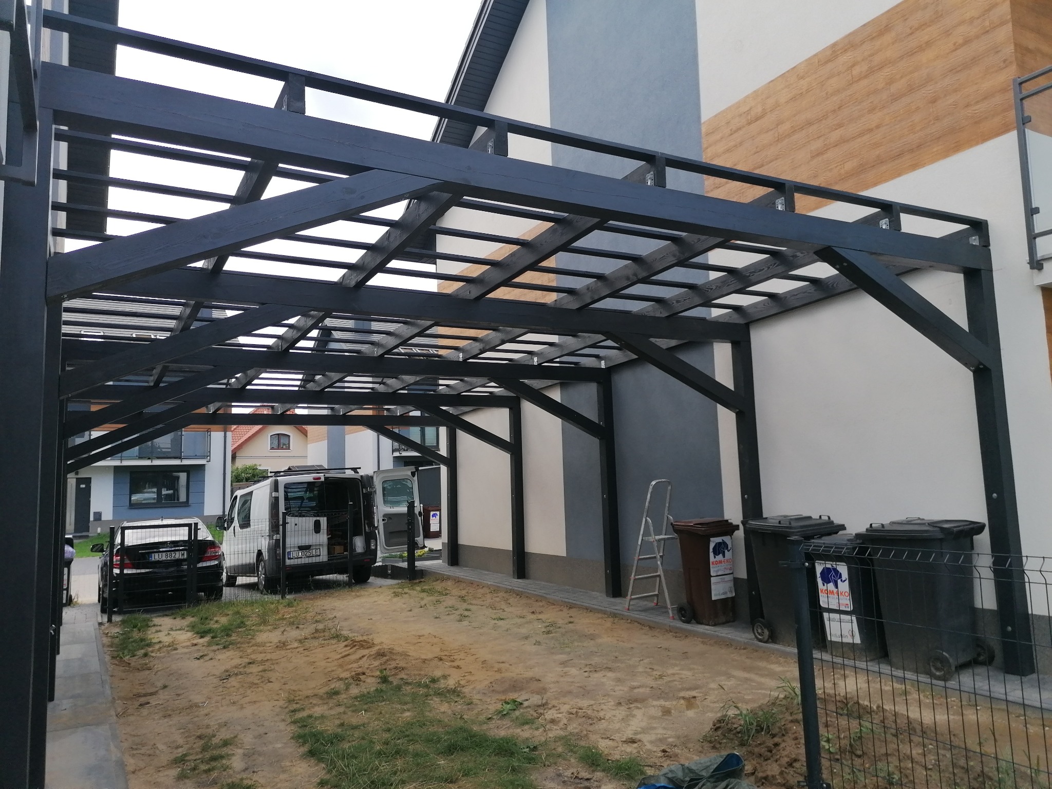 Czarna drewniana konstrukcja zadaszenia na posesji, samochody i kontenery na śmieci w tle, widoczna drabina oparta o ścianę budynku.