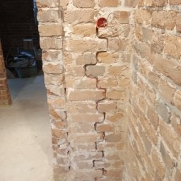 Piotr Milke - Wykuta bruzda w ceglanej ścianie pod instalację elektryczną, widoczne przewody i puszka elektryczna, w tle surowy stan pomieszczenia w trakcie remontu.