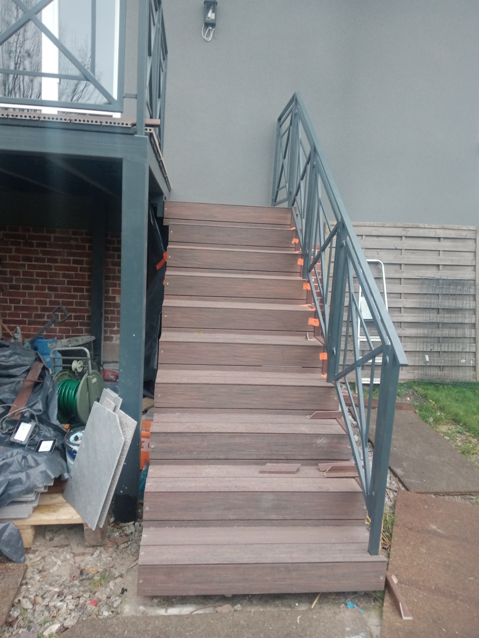 Zewnętrzne schody z kompozytu drewna z metalową balustradą w trakcie montażu, widoczne pomarańczowe elementy mocujące, narzędzia i materiały budowlane w tle.