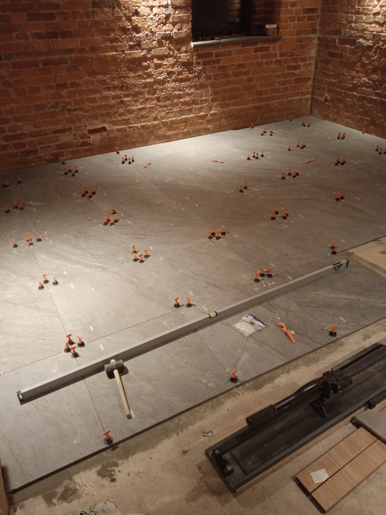 Układanie szarych płytek na podłodze z użyciem poziomicy, młotka gumowego i klipsów poziomujących, widoczna surowa ściana z czerwonej cegły.