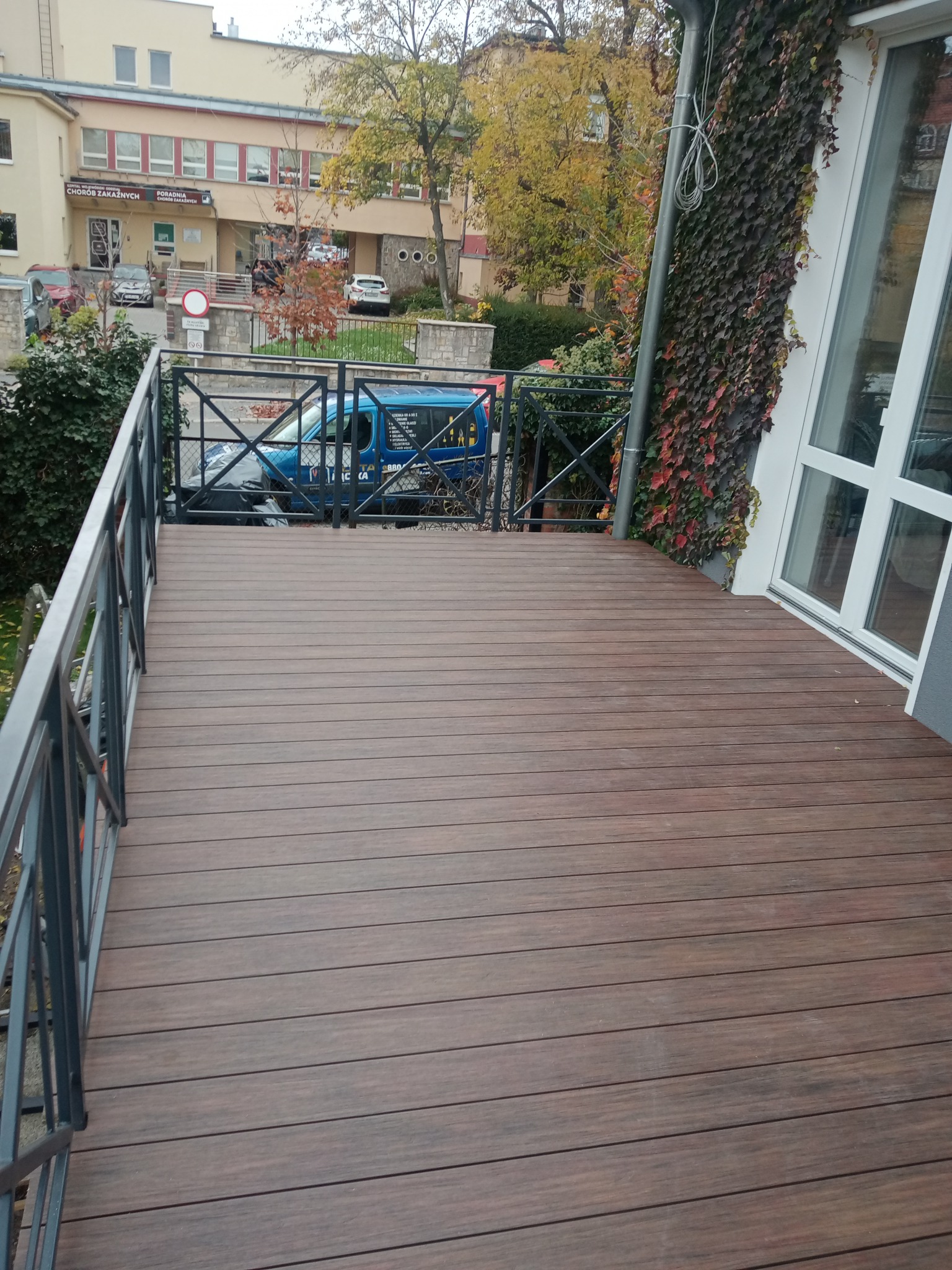 Nowy taras z desek kompozytowych w kolorze ciemnego drewna, otoczony metalową balustradą, z widokiem na budynek i zaparkowany samochód z narzędziami.