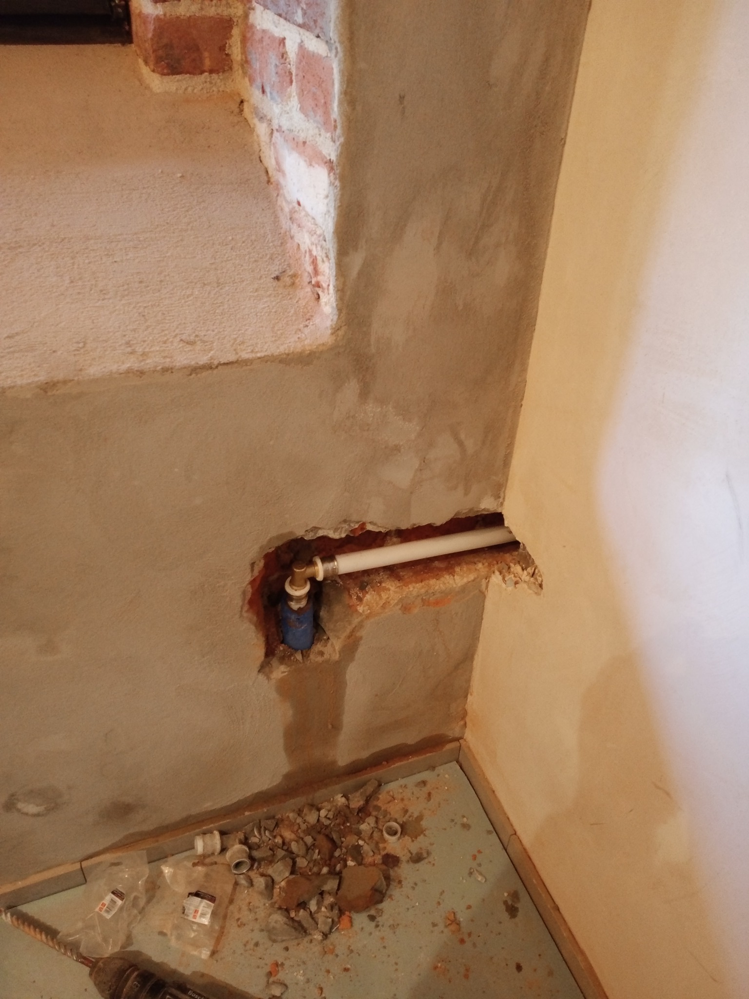 Fragment ściany z odsłoniętymi rurami instalacji wodnej w trakcie prac remontowych, widoczne gruz i narzędzia na podłodze, surowy stan wykończenia.