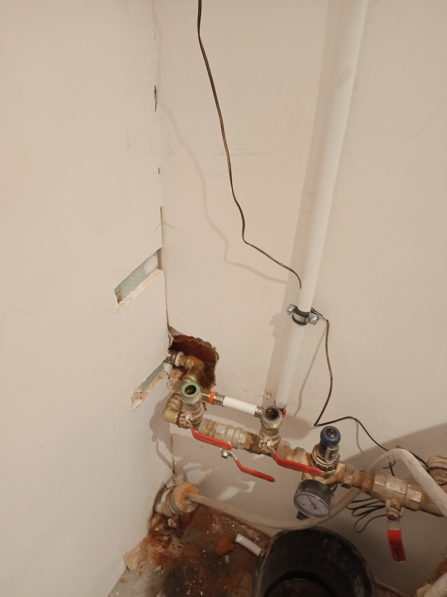 Fragment ściany podczas prac instalacyjnych z widocznymi rurami, zaworami, manometrem i otworami w ścianie, w tle biała rura pionowa przymocowana obejmą.