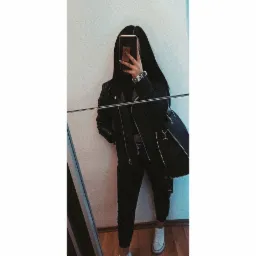 Osoba w czarnej kurtce i spodniach pozuje do selfie w lustrze, zasłaniając twarz telefonem, widoczna torebka z ćwiekami i białe trampki.