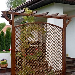 REKWIZYTORNIA KRZYSZTOF HULBÓJ - Drewniana pergola z kratką, częściowo porośnięta bluszczem, na tle białej ściany domu z brązowym dachem, z widocznym doniczką z kwiatami po lewej stronie.
