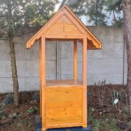 REKWIZYTORNIA KRZYSZTOF HULBÓJ - Drewniana konstrukcja z daszkiem i wyciętym krzyżem, prawdopodobnie studzienka, na tle betonowego ogrodzenia i drzew