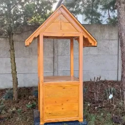 Drewniana konstrukcja z daszkiem i wyciętym krzyżem, prawdopodobnie studzienka, na tle betonowego ogrodzenia i drzew