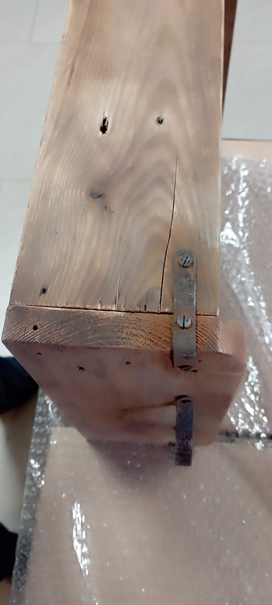 Fragment surowego, drewnianego blatu z widocznymi sękami, pęknięciami i metalowymi mocowaniami, leżący na folii bąbelkowej.