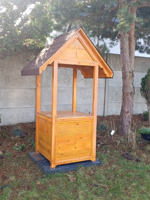 Drewniana kapliczka ogrodowa z brązowym dachem, widok z przodu na tle zieleni i betonowego ogrodzenia.