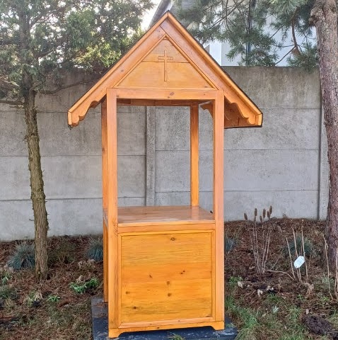 Drewniana konstrukcja z daszkiem i wyciętym krzyżem, prawdopodobnie studzienka, na tle betonowego ogrodzenia i drzew