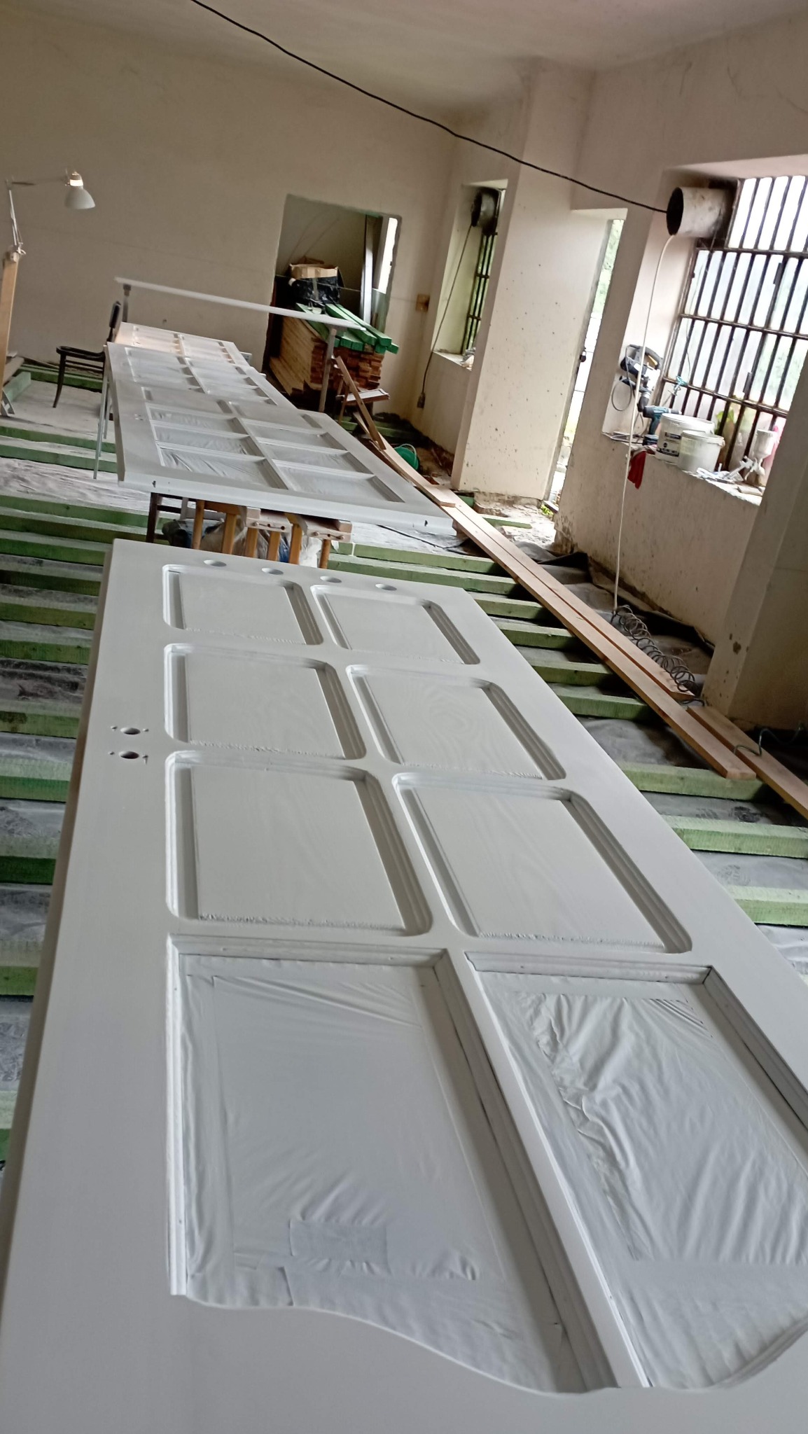 Białe, drewniane drzwi z panelami zabezpieczonymi folią malarską, leżące na podkładkach w trakcie renowacji w jasnym pomieszczeniu.