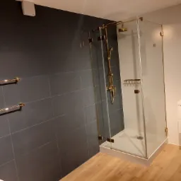 Nowoczesna łazienka z kabiną prysznicową ze złotymi elementami, ciemnoszare płytki na ścianie, drewnopodobna podłoga i biała umywalka z szafką.