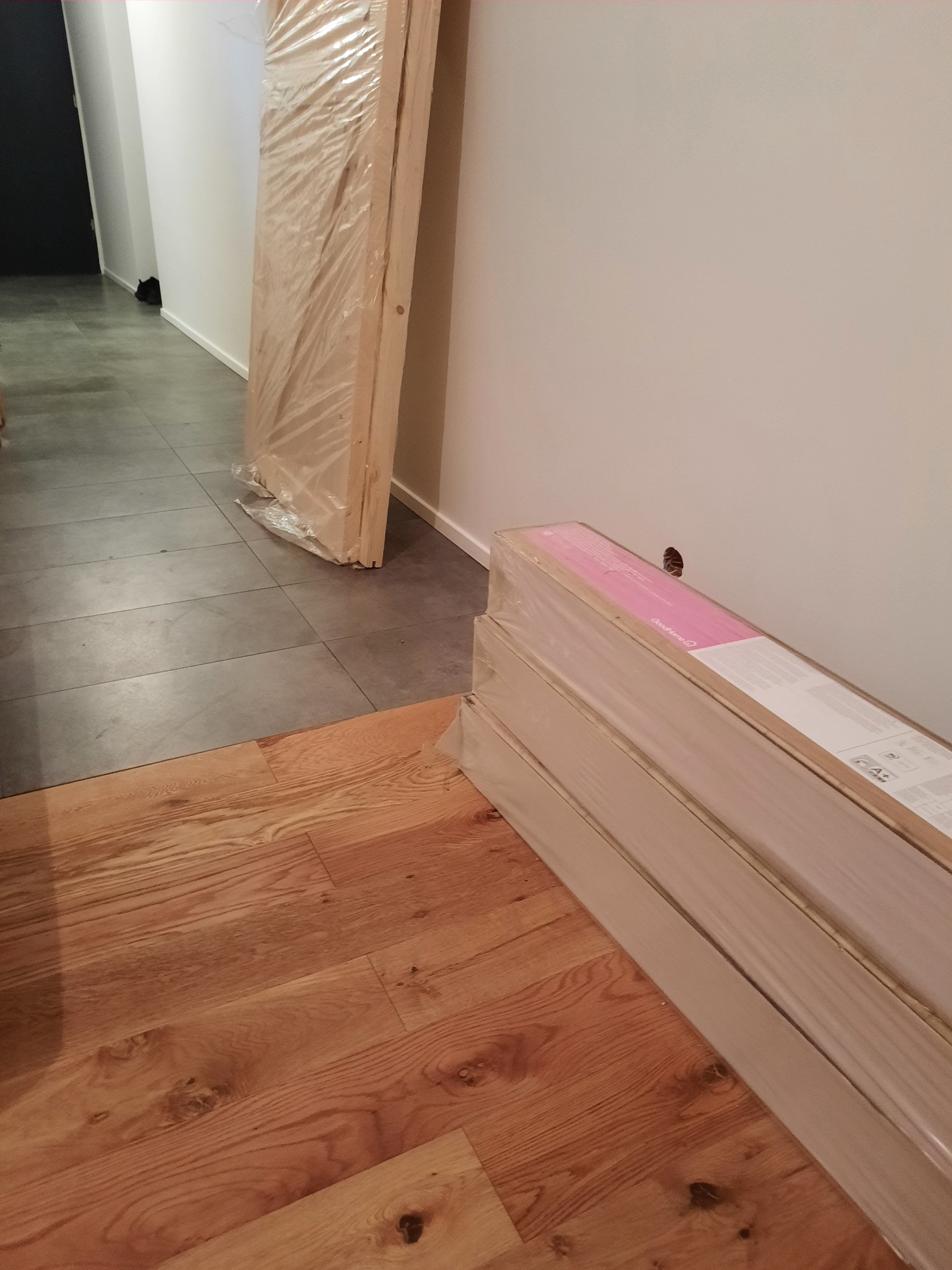 Paczki paneli podłogowych GoodHome w folii, oraz drzwi w folii oparte o ścianę w korytarzu z wyłożoną podłogą z płytek ceramicznych, fragment ułożonej drewnianej podłogi.