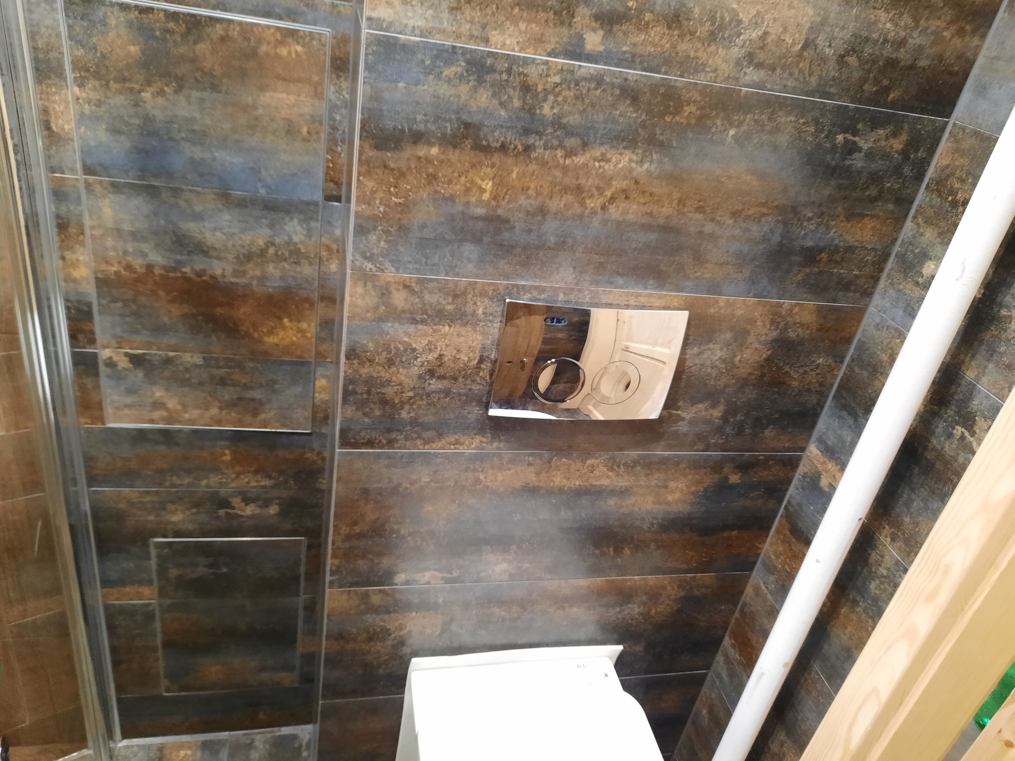 Nowoczesna łazienka z brązowo-szarymi płytkami imitującymi drewno, wisząca toaleta z chromowanym przyciskiem spłukującym i fragment kabiny prysznicowej.