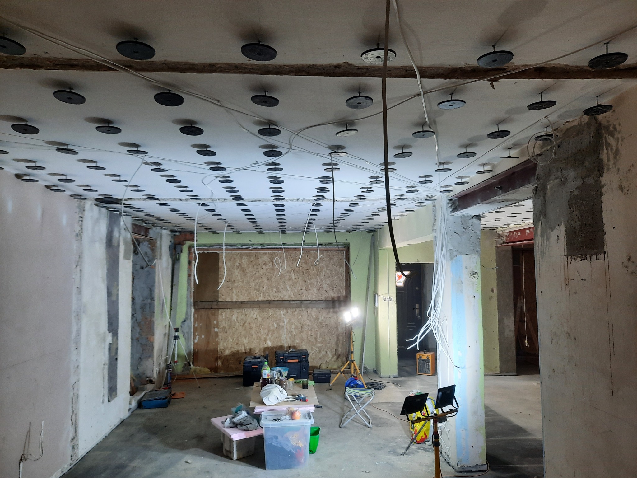 Wnętrze w trakcie prac przygotowawczych do sufitu podwieszanego, widoczne zamocowane uchwyty, przewody elektryczne oraz narzędzia i materiały budowlane na prowizorycznym stole.