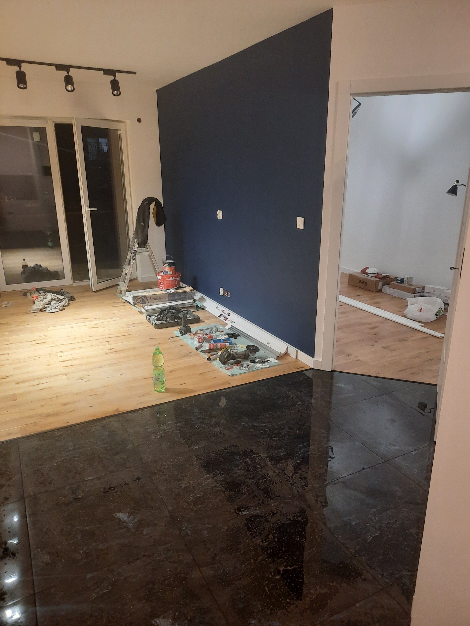 Wnętrze w trakcie prac wykończeniowych; widoczna świeżo pomalowana na granatowo ściana, częściowo ułożona podłoga z paneli przechodząca w ciemne płytki, rozstawiona drabina z narzędziami i puszkami...